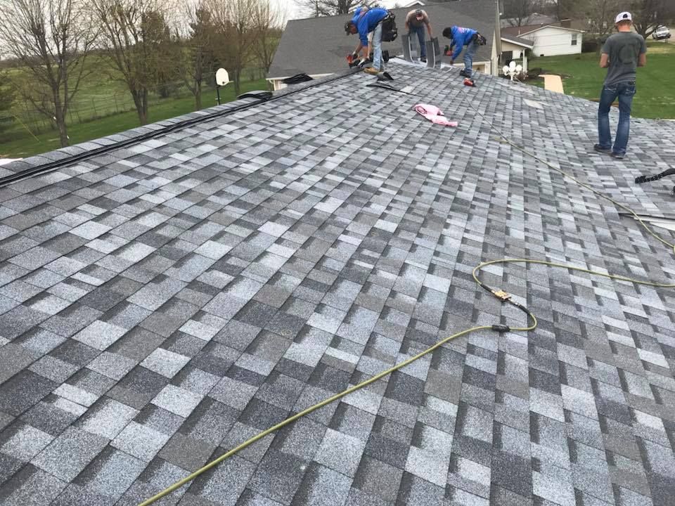 Workers installing gray asphalt shingles on a steep roof outdoors.