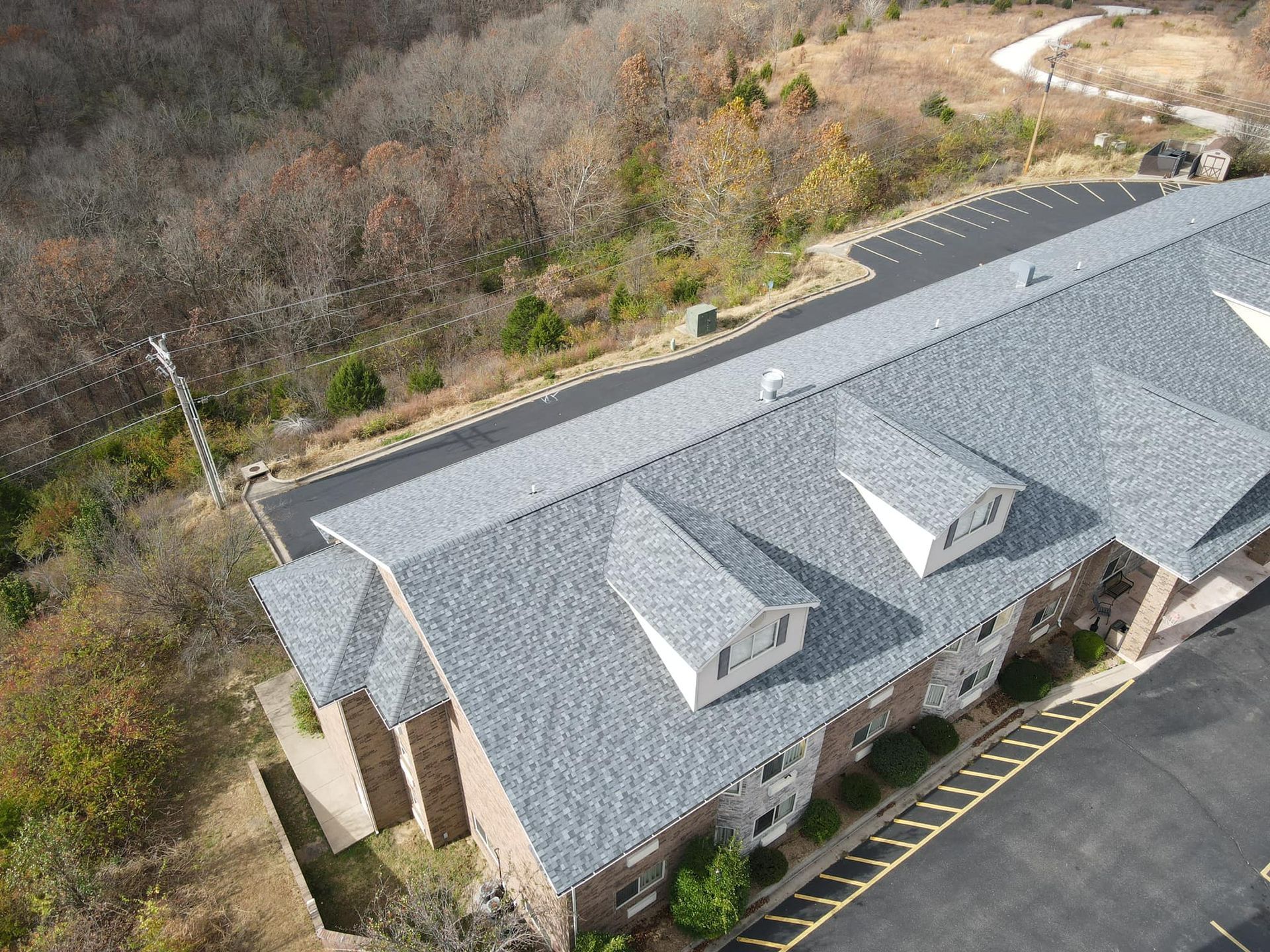 Aerial view of a gray-roofed building beside a parking lot and wooded hillside.