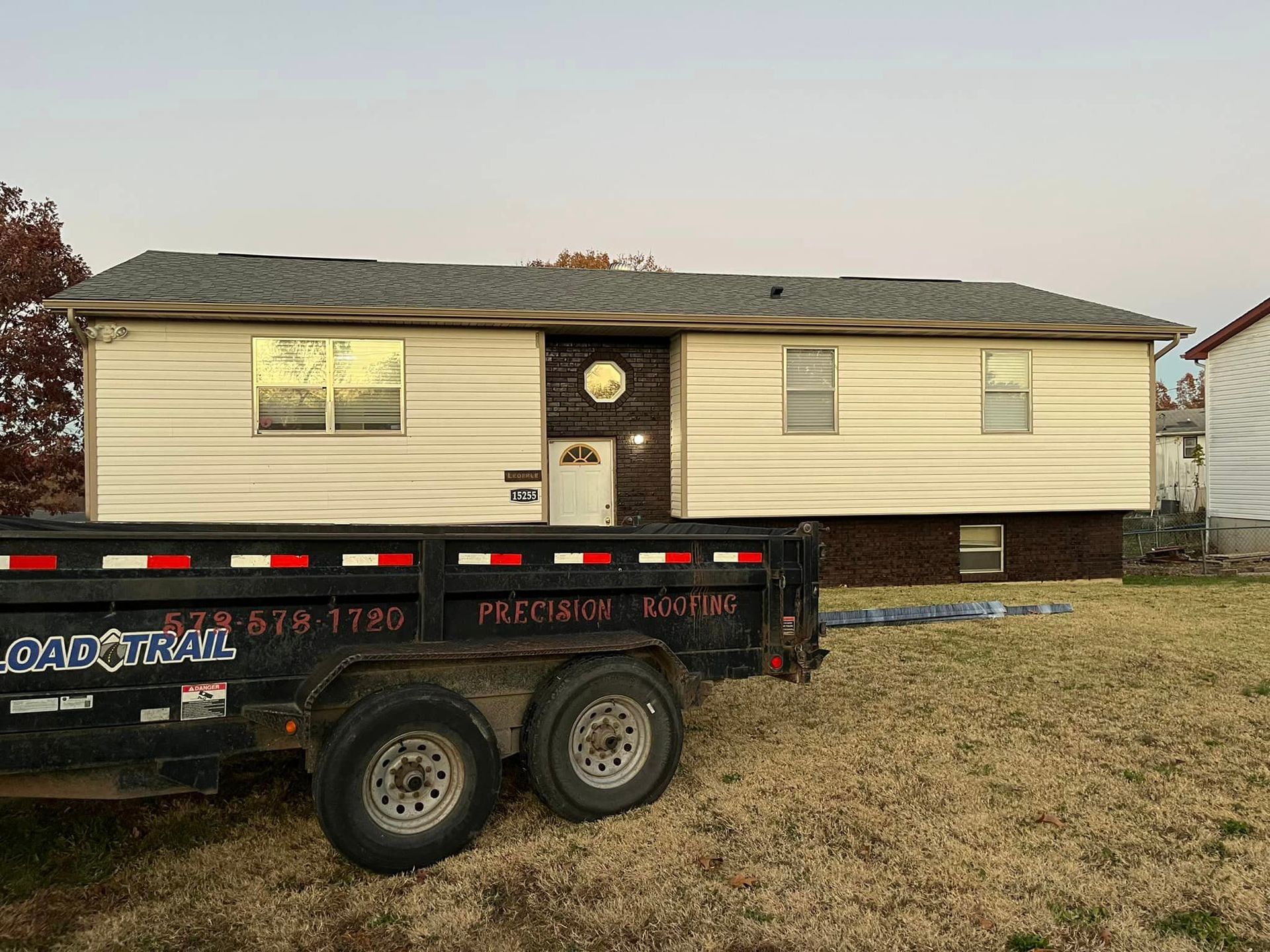 Single-wide beige mobile home behind a black dump trailer on a grassy lot.