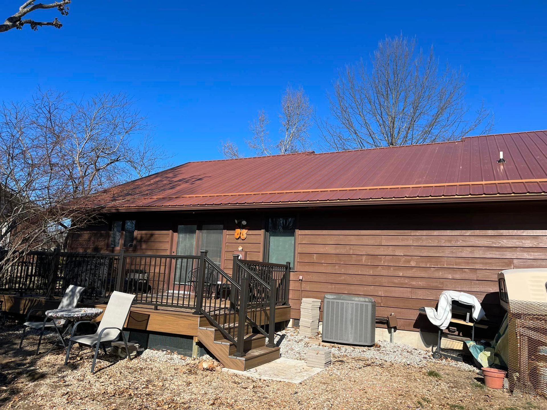 Brown house with a rust-red metal roof, small front porch, and outdoor chairs under a clear blue sky
