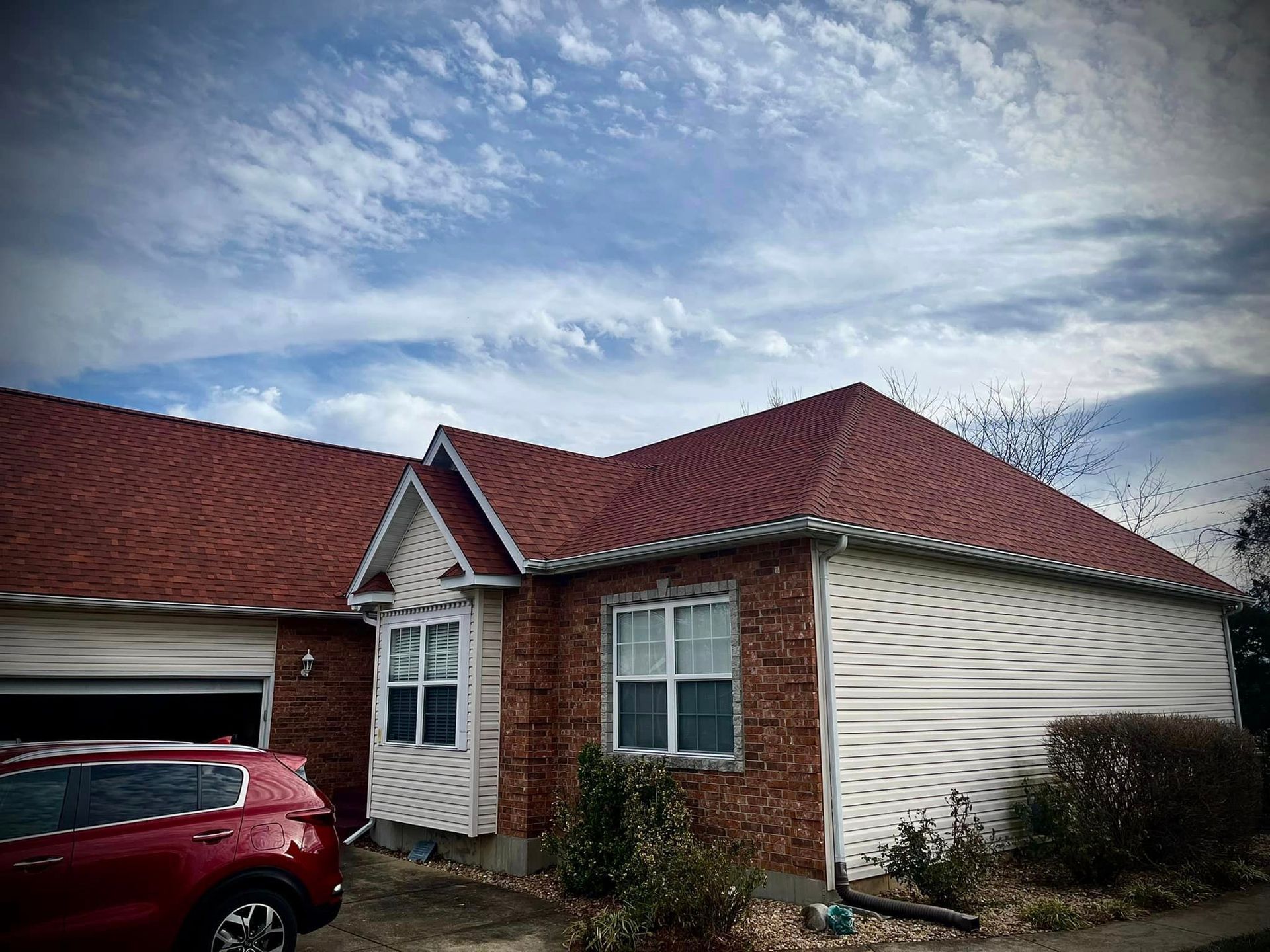 Single-story brick house with red roof and a red SUV parked in the driveway under a cloudy sky