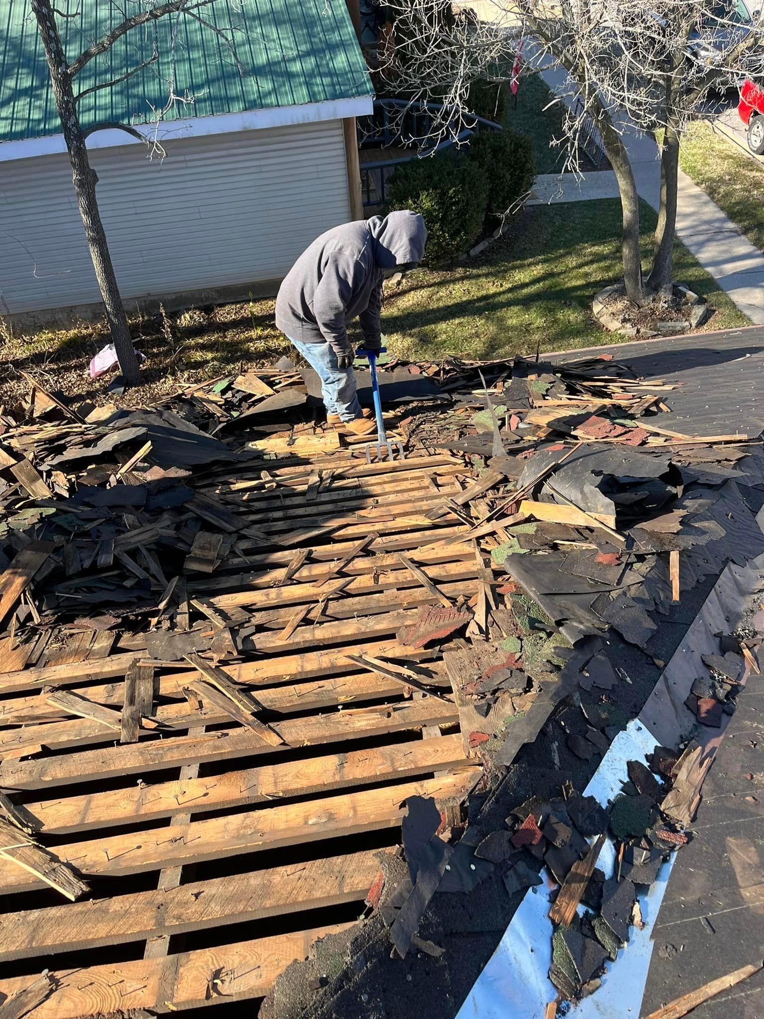 Person in a hoodie clearing debris from a damaged roof with exposed wooden beams.