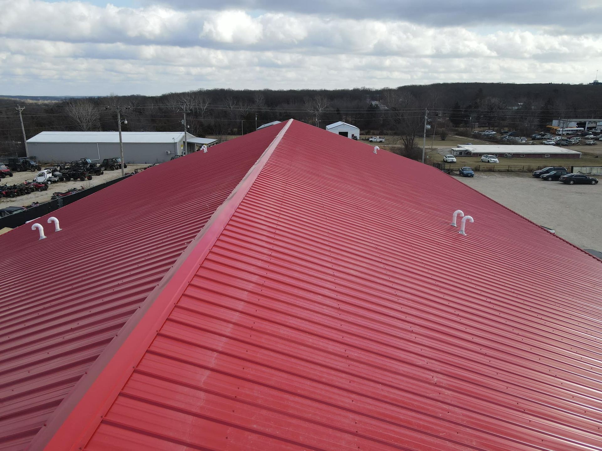 Red metal roof ridge with rows of seams, viewed from above over a cloudy industrial landscape.