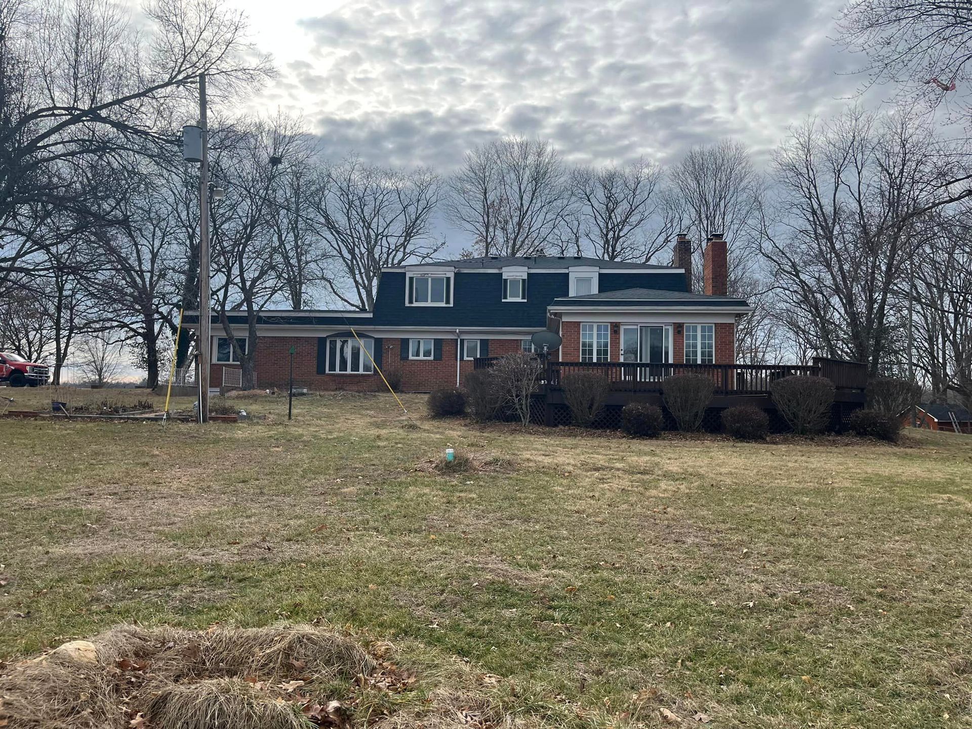 Brick house with dark roof and white porch on a grassy lot under a cloudy sky, framed by leafless trees