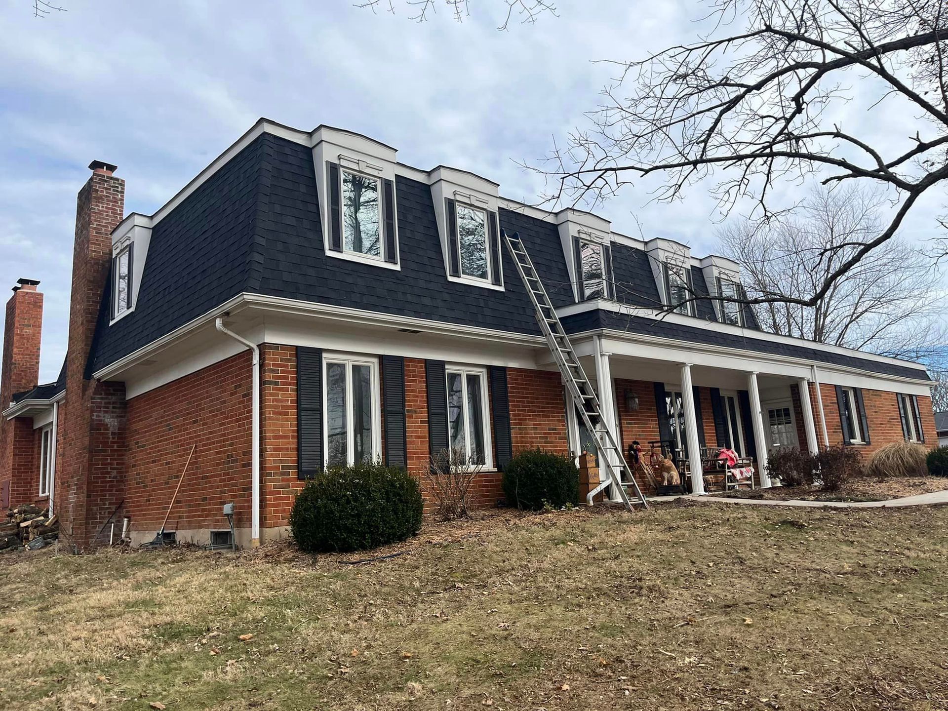 Large brick house with dark mansard roof and white columns on a sloping lawn under a gray sky