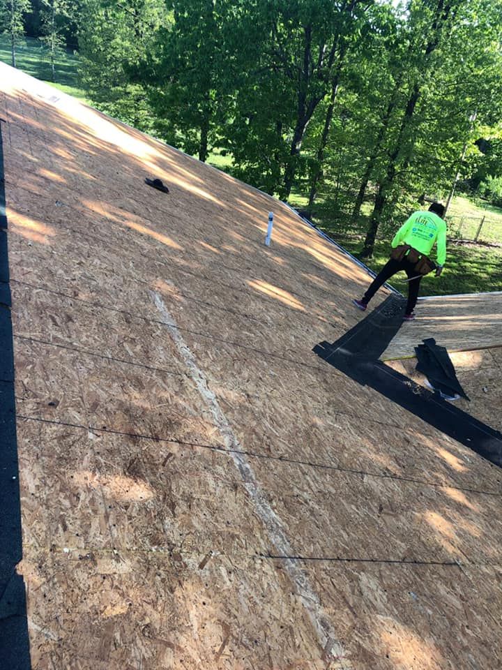 Worker in neon green shirt on a steep roof, applying roofing material in a wooded area.