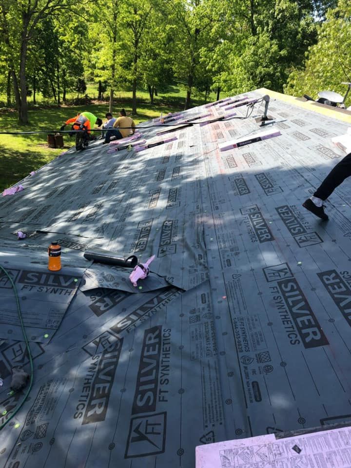 Workers laying roofing underlayment on a steep roof in a wooded yard, with tools nearby.