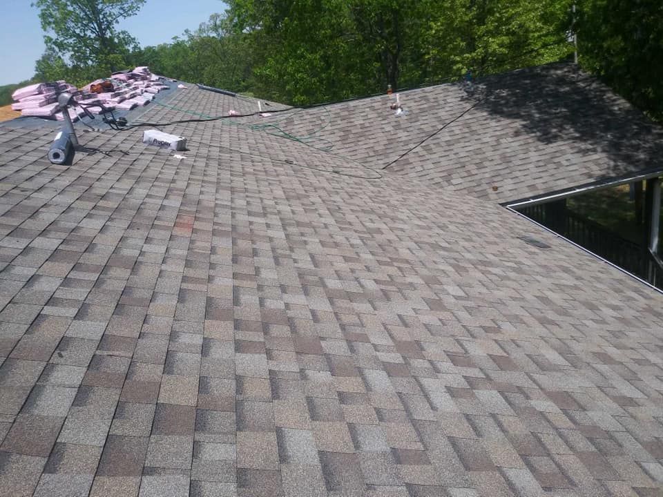 Roofers installing shingles on a steep gray roof, with bundles of materials near the ridge and trees in the background.