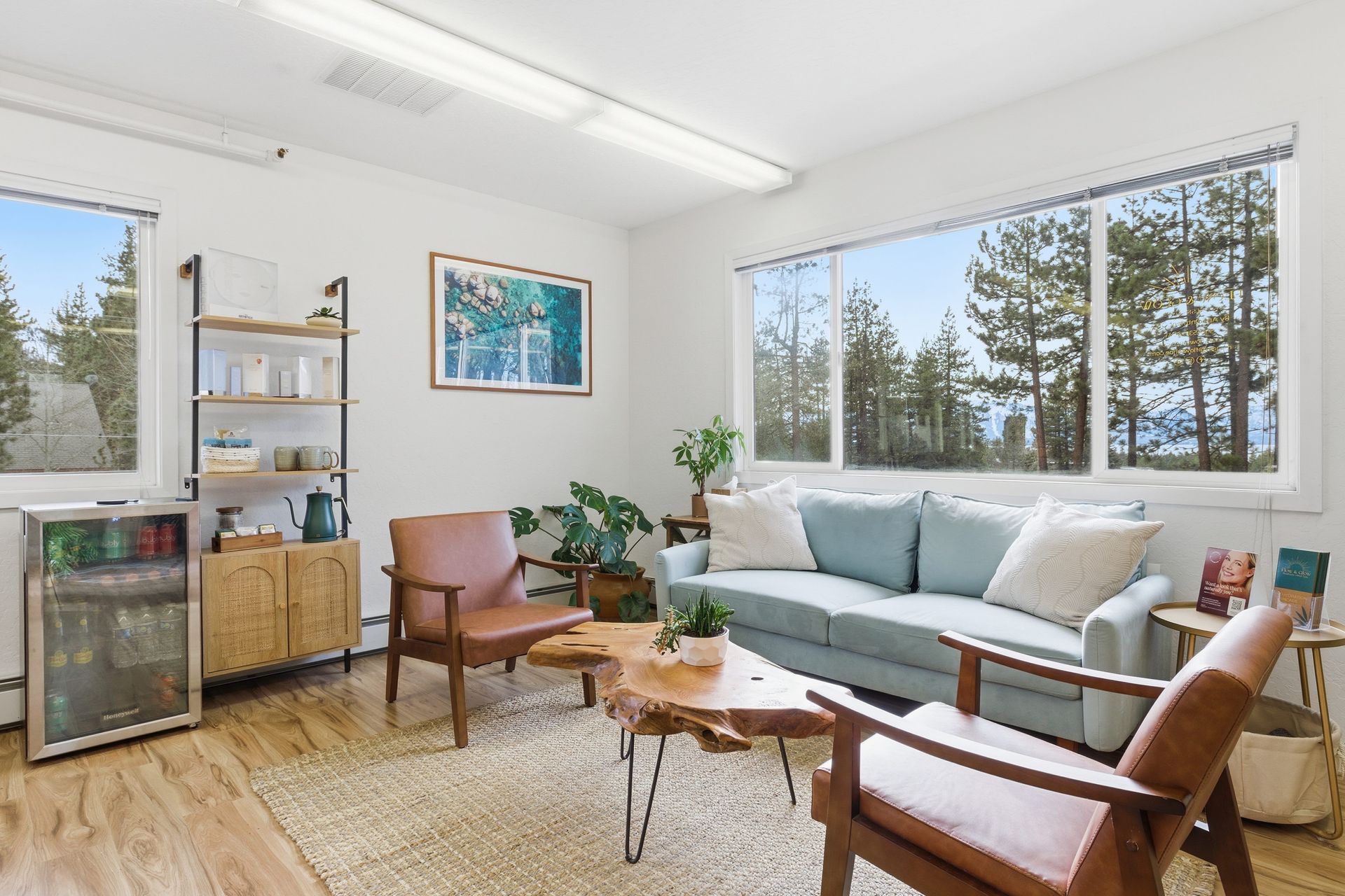 Bright living room with blue sofa, wooden chairs, coffee table, shelving, and large windows overlooking trees