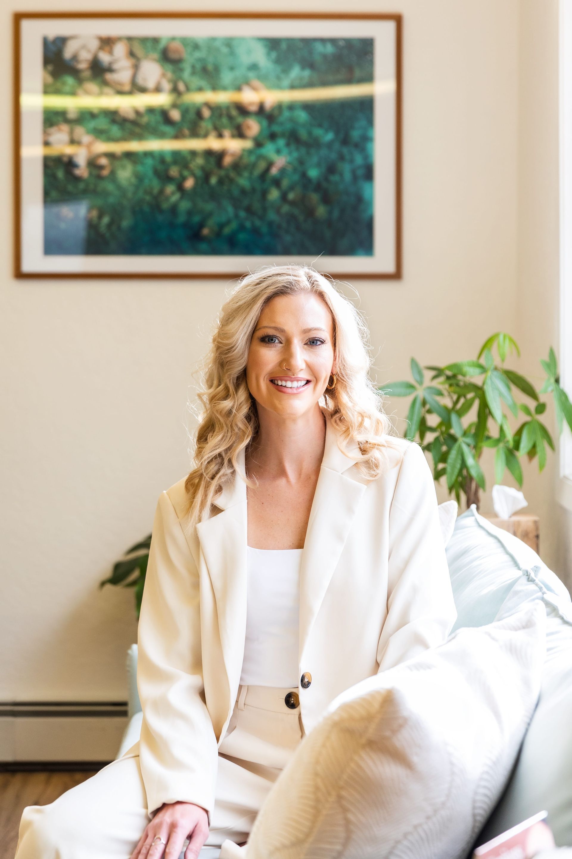Smiling person seated on a couch in a bright living room with a framed landscape and a potted plant.