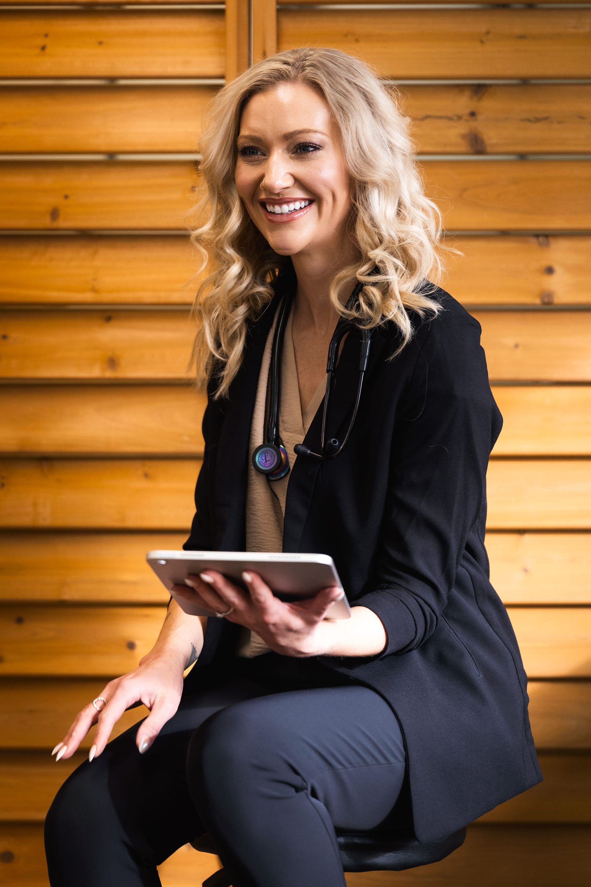 Smiling woman in black outfit sitting on a bench, holding a tablet against a wooden wall.