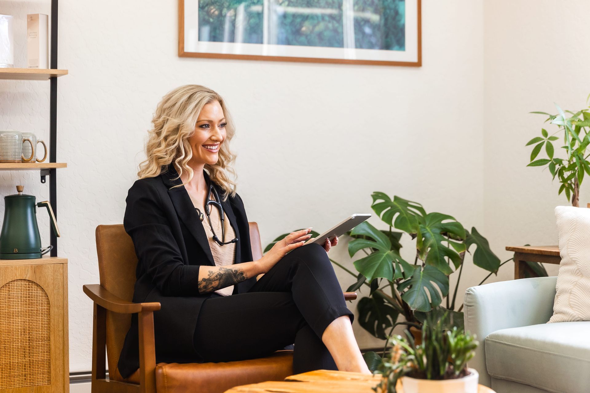 Woman sitting in a cozy office lounge, smiling and using a tablet beside a plant and sofa.