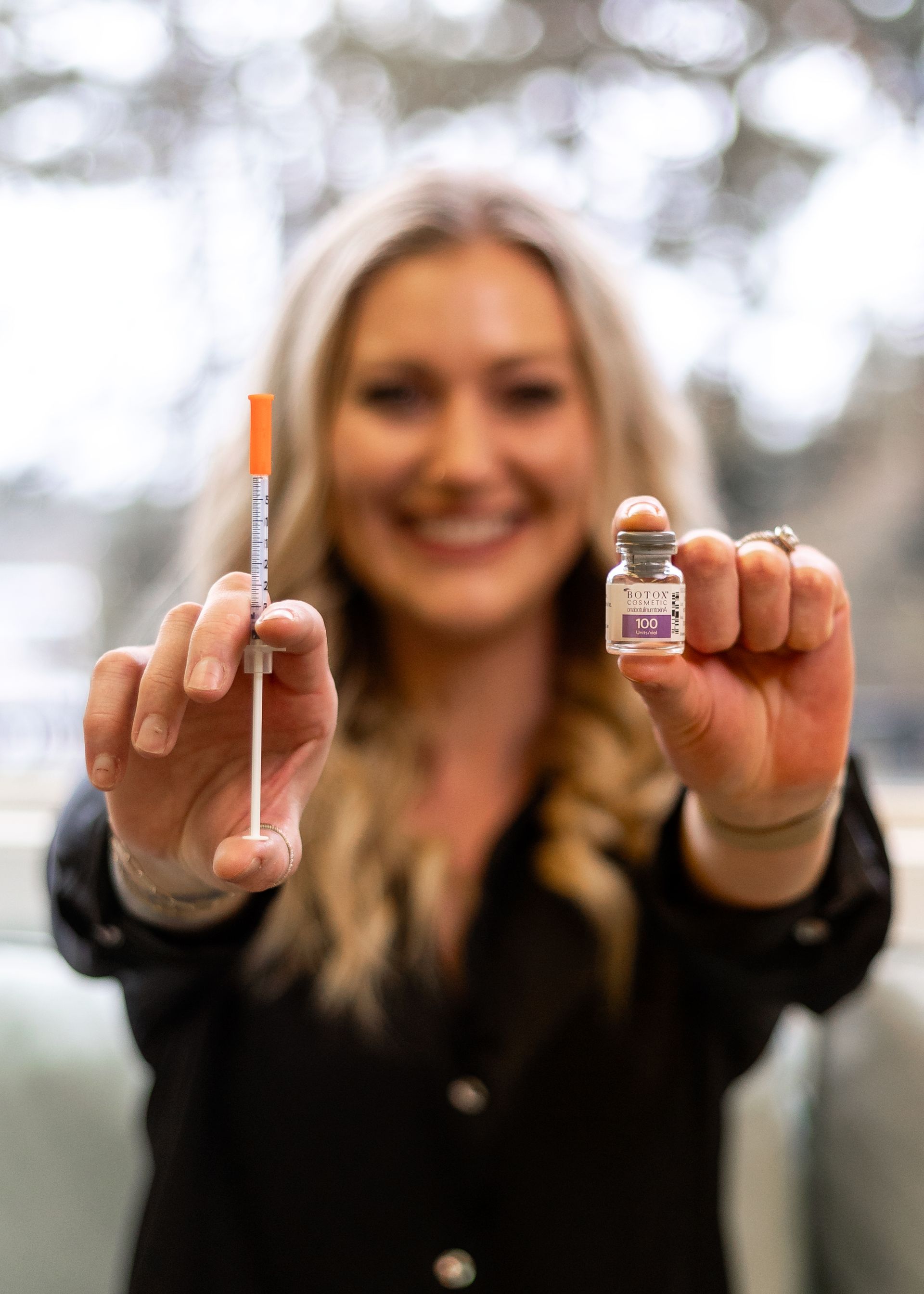 Person holding a syringe and a small vial toward the camera, smiling outdoors.