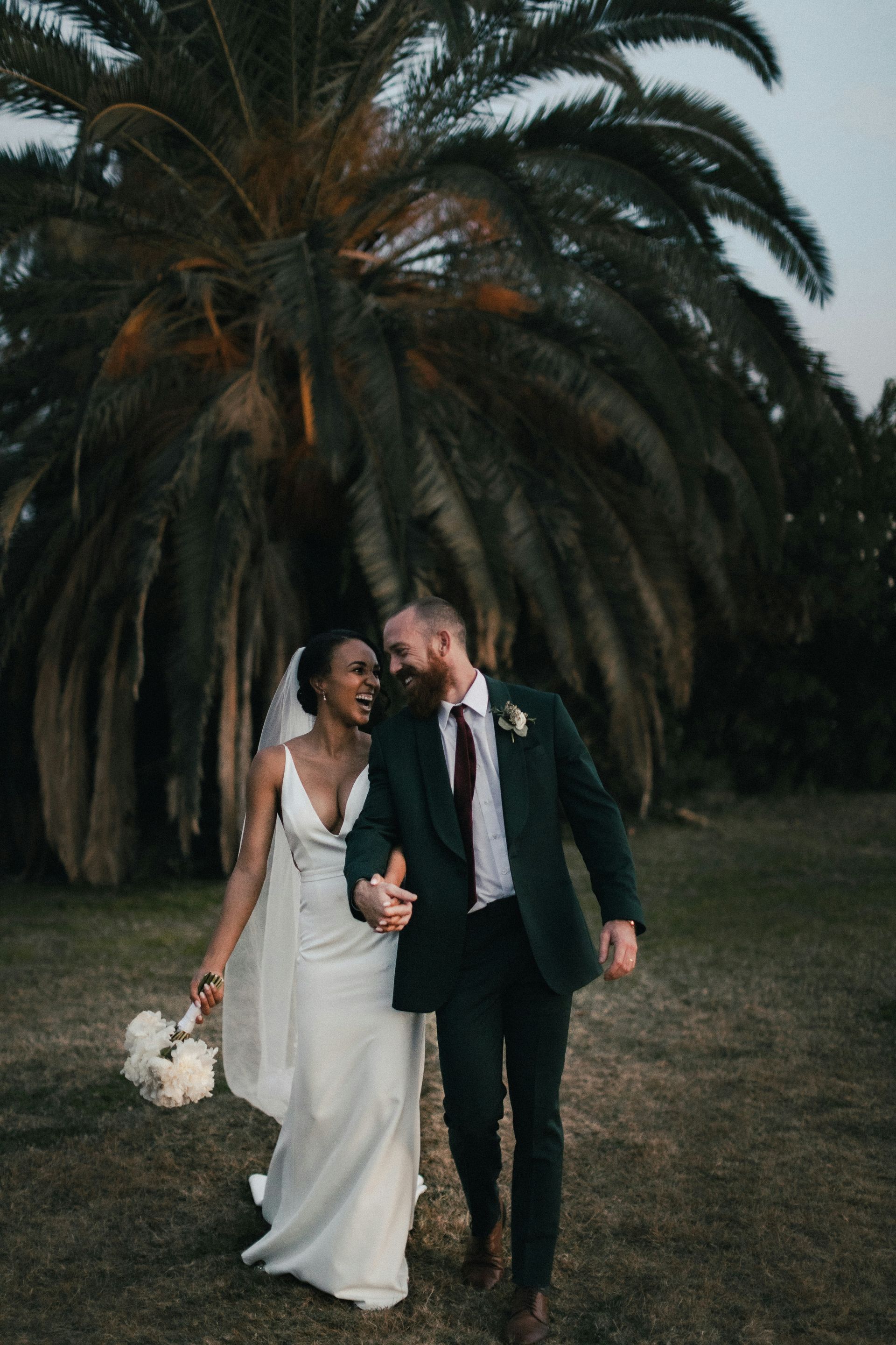 Bride and groom walking arm in arm in a garden, holding a bouquet before a large palm tree
