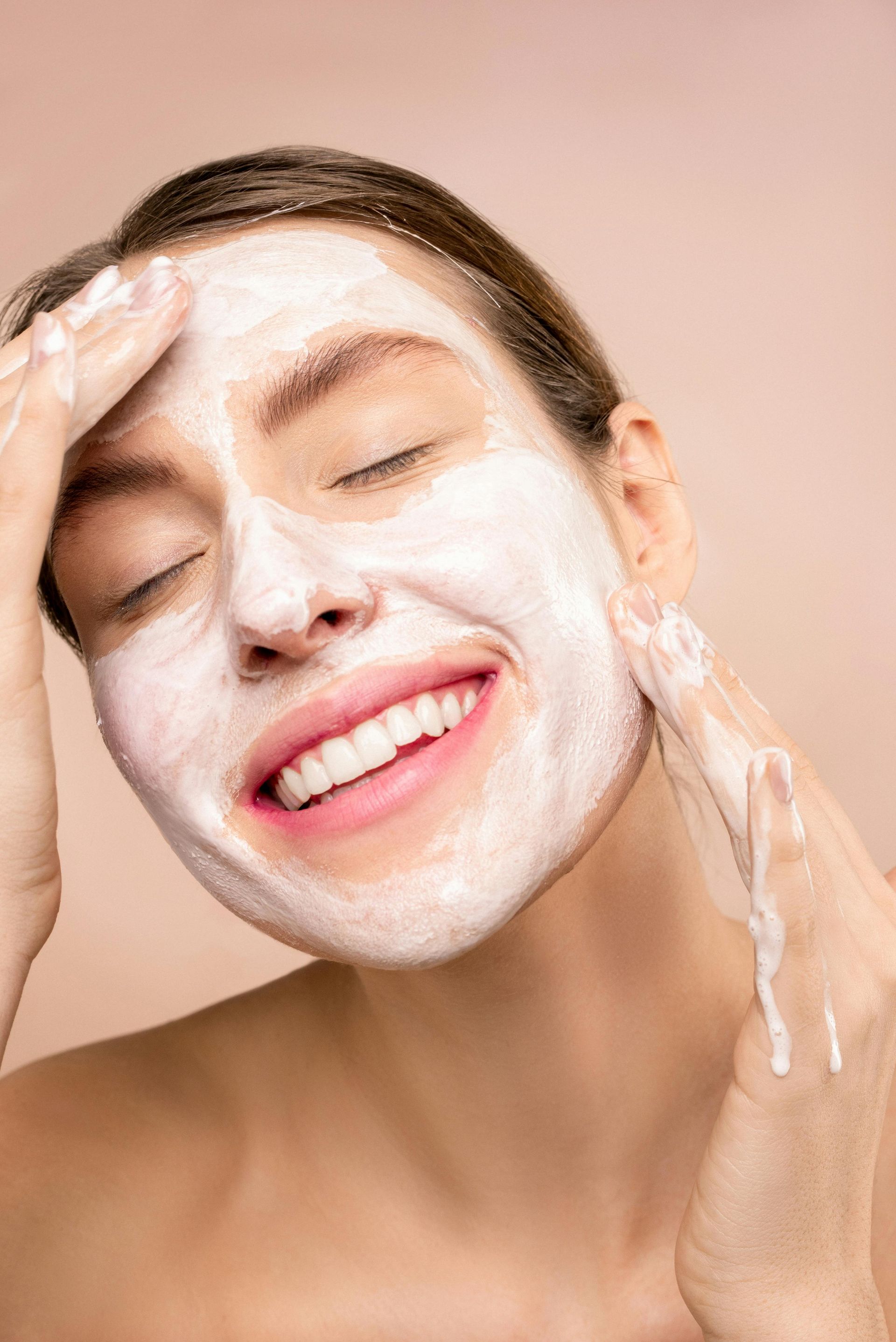 Person applying white facial cleanser and smiling with eyes closed against a pink background