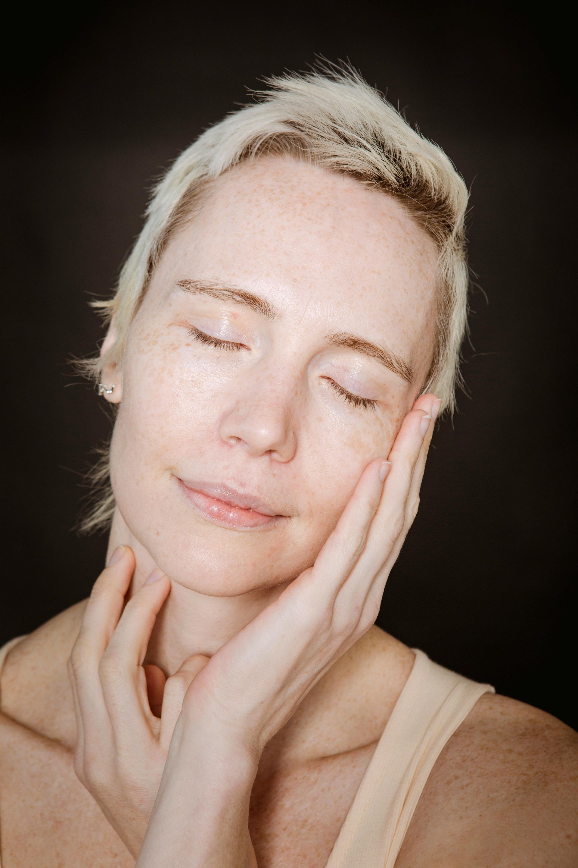 Close-up of a person with closed eyes, gently touching their face against a dark background