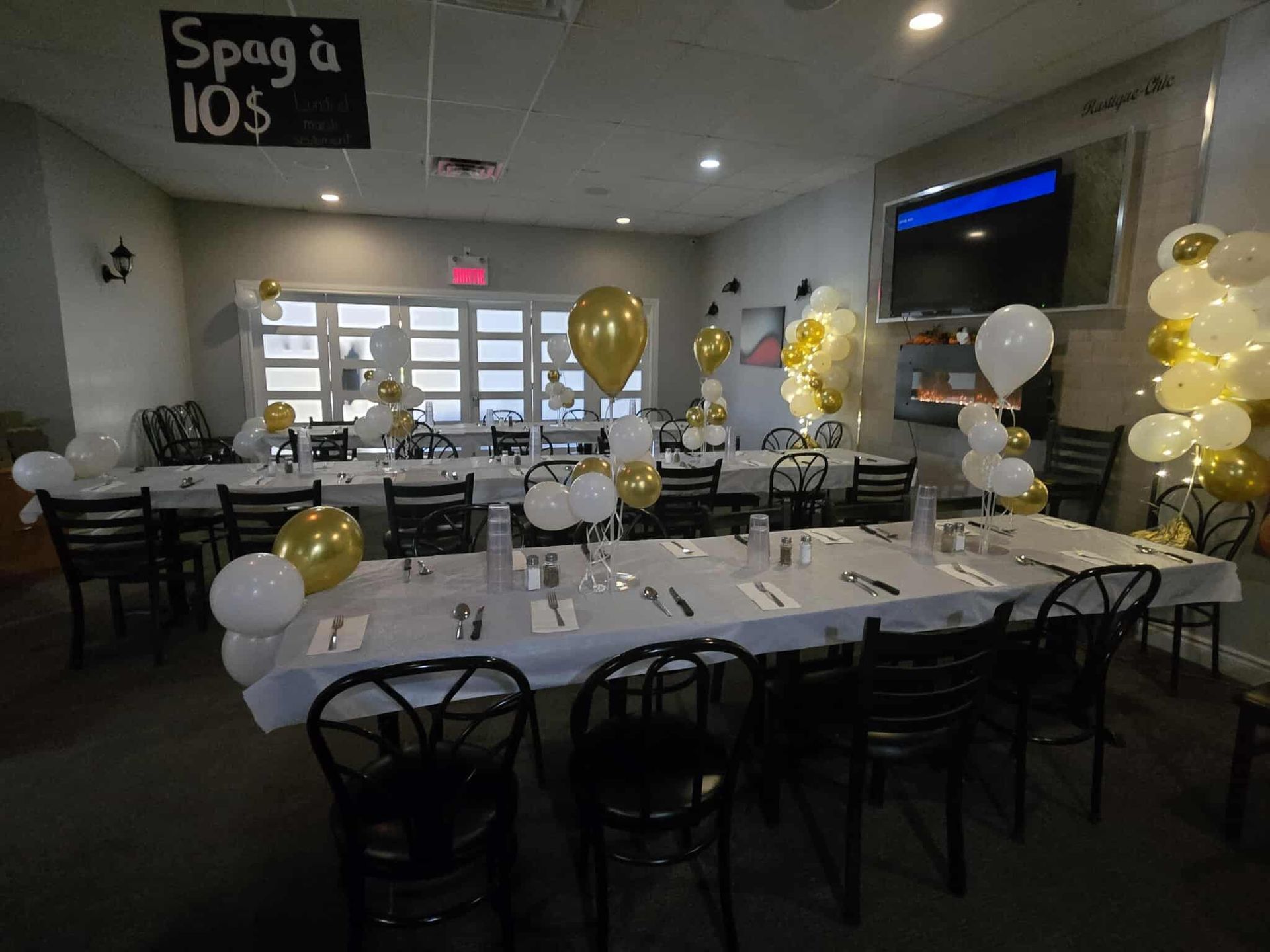 Une salle à manger dressée pour une fête, avec des tables nappées de blanc, des chaises noires et des grappes de ballons dorés et blancs.