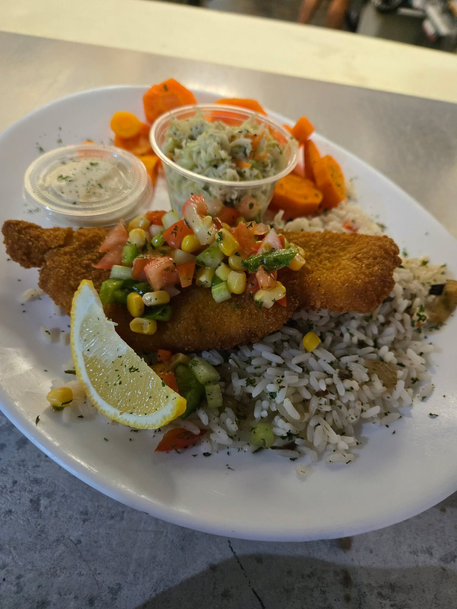 Une assiette de poisson frit nappée de salsa de maïs, servie avec du riz, des carottes coupées en rondelles et une sauce, accompagnée d'un quartier de citron.
