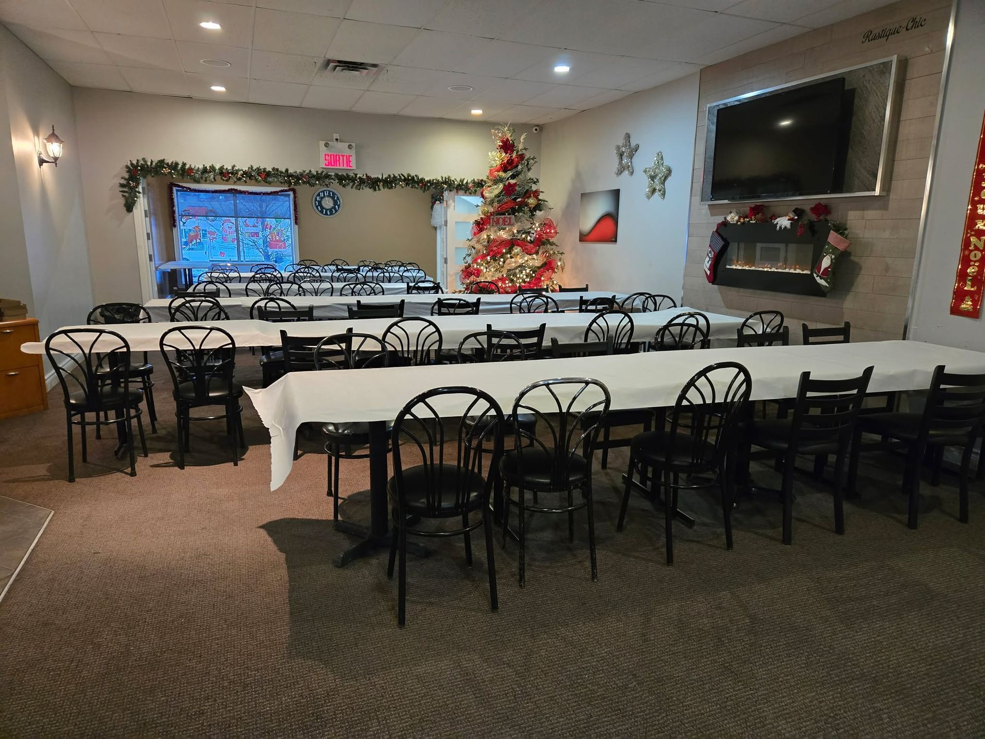 Une salle de banquet aménagée pour une fête de fin d'année, avec des rangées de tables nappées de blanc, des chaises noires et un sapin de Noël décoré.