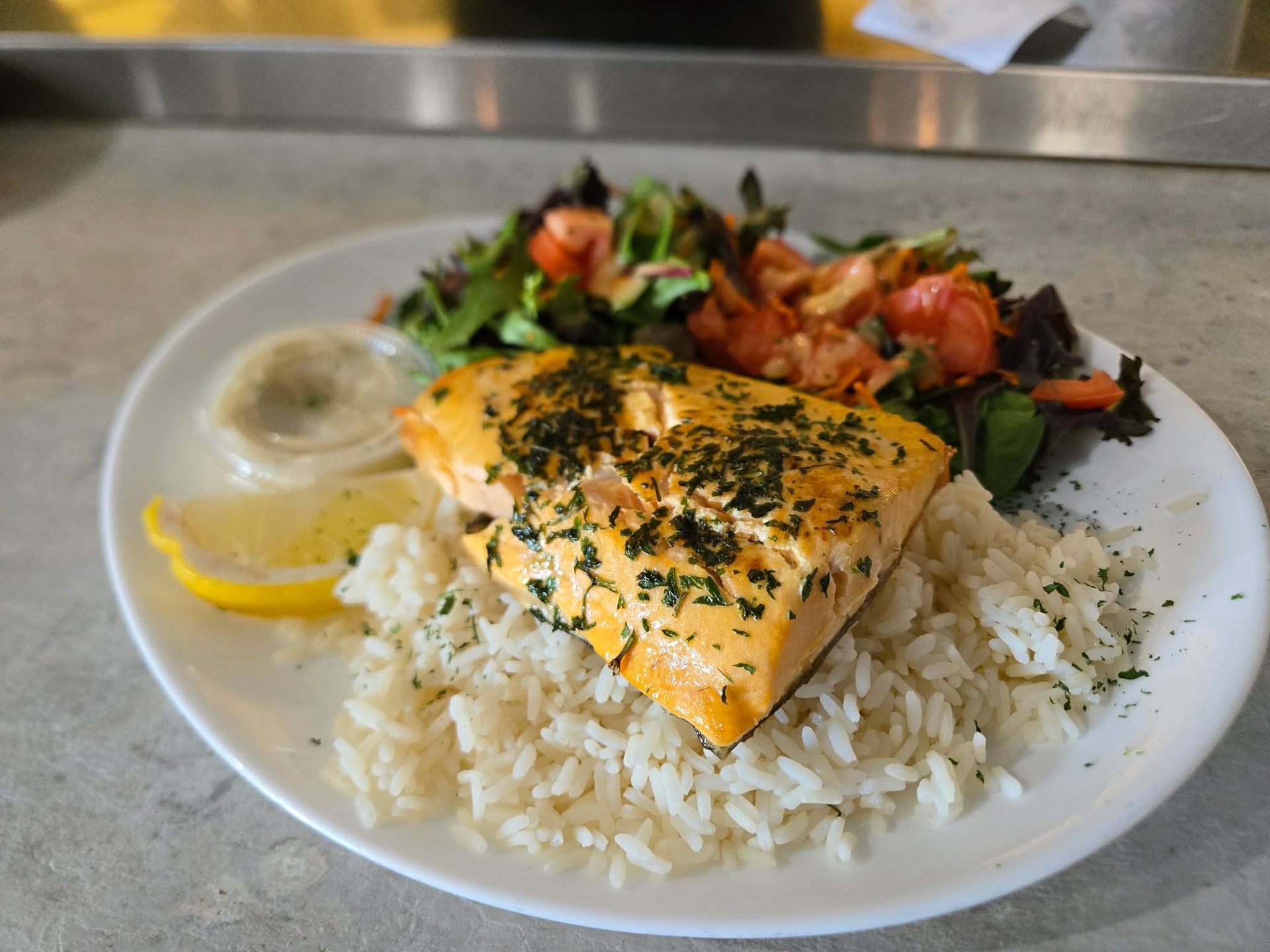Une assiette de saumon cuit garnie d'herbes, servie sur du riz blanc avec une salade d'accompagnement, un quartier de citron et une sauce.