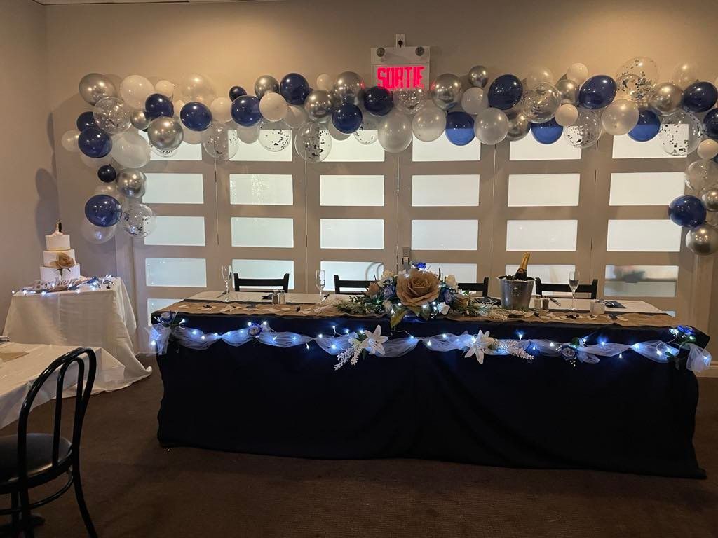 Une table de banquet festive décorée de ballons bleus et blancs, de guirlandes lumineuses et d'un gâteau blanc sur une table d'appoint.