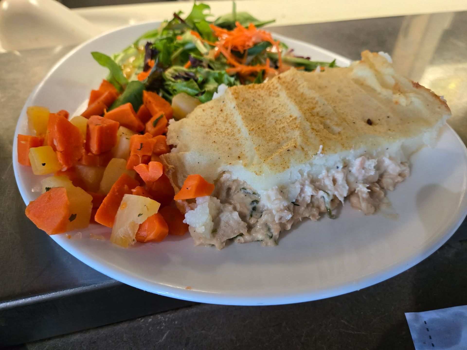 Une assiette composée d'une portion de tourte au poisson recouverte de purée de pommes de terre, accompagnée de carottes sautées et d'une salade verte.