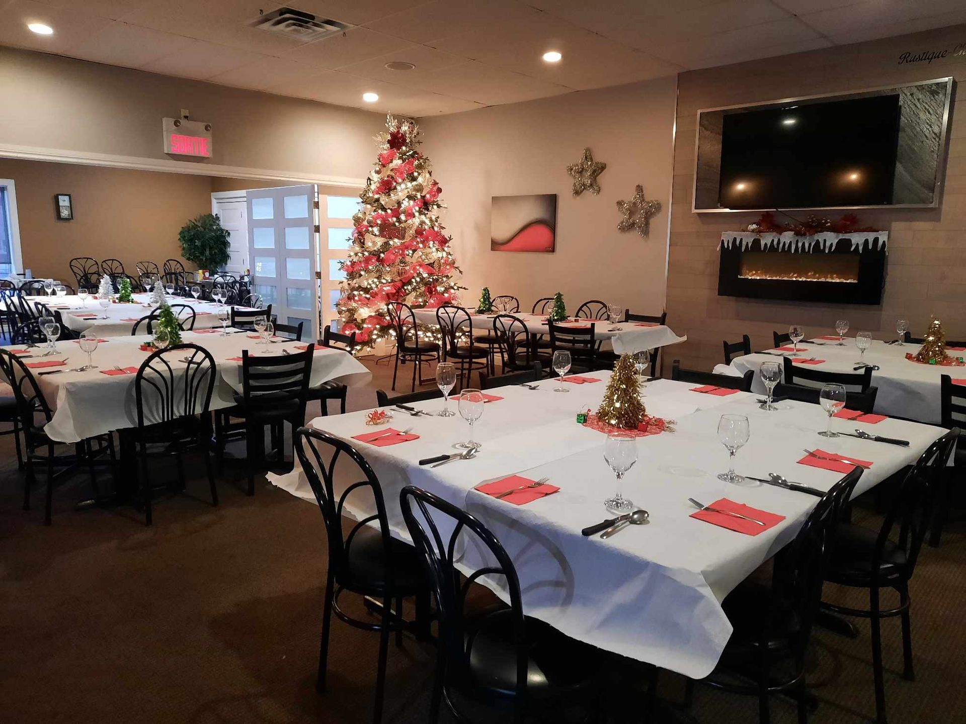 Une salle à manger de restaurant décorée pour Noël, avec des tables recouvertes de nappes blanches, de serviettes rouges et de petits sapins.