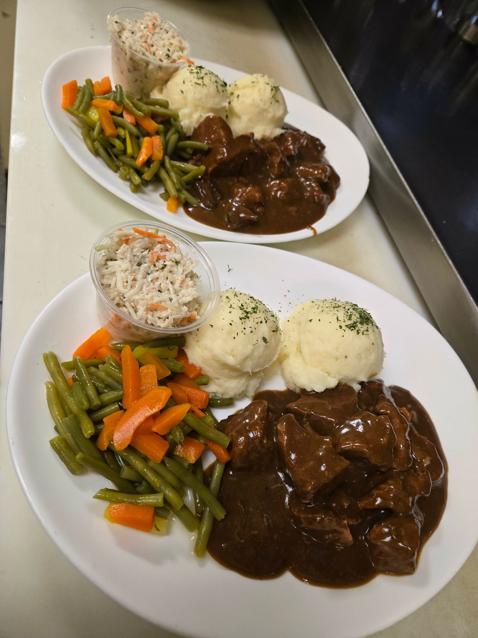 Deux assiettes ovales blanches, chacune servie avec du ragoût de bœuf, de la purée de pommes de terre, un mélange de légumes composé de haricots verts et de carottes, et de la salade de chou.