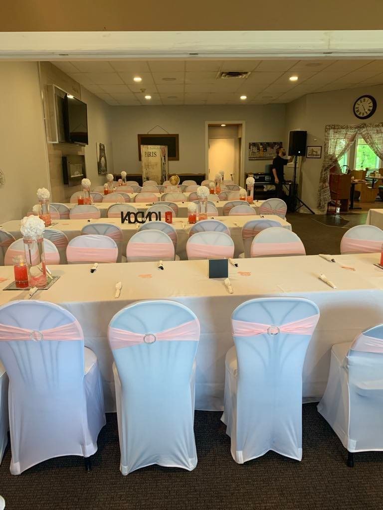 Une salle de banquet avec des rangées de tables dressées pour un événement, avec des housses de chaises blanches ornées de rubans rose poudré.