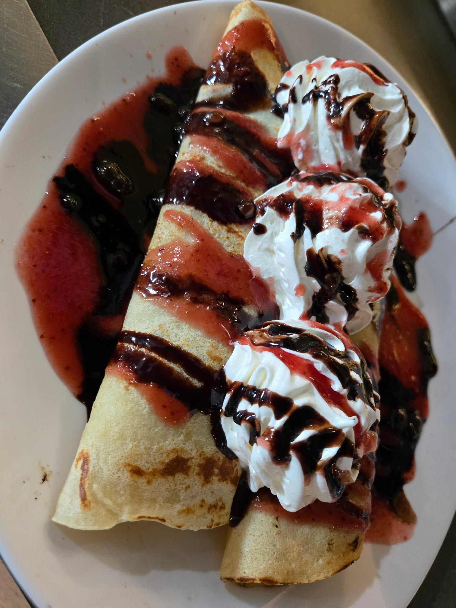 Deux crêpes roulées sur une assiette, garnies de trois quenelles de crème fouettée et arrosées de sirop de chocolat et de fruits rouges.