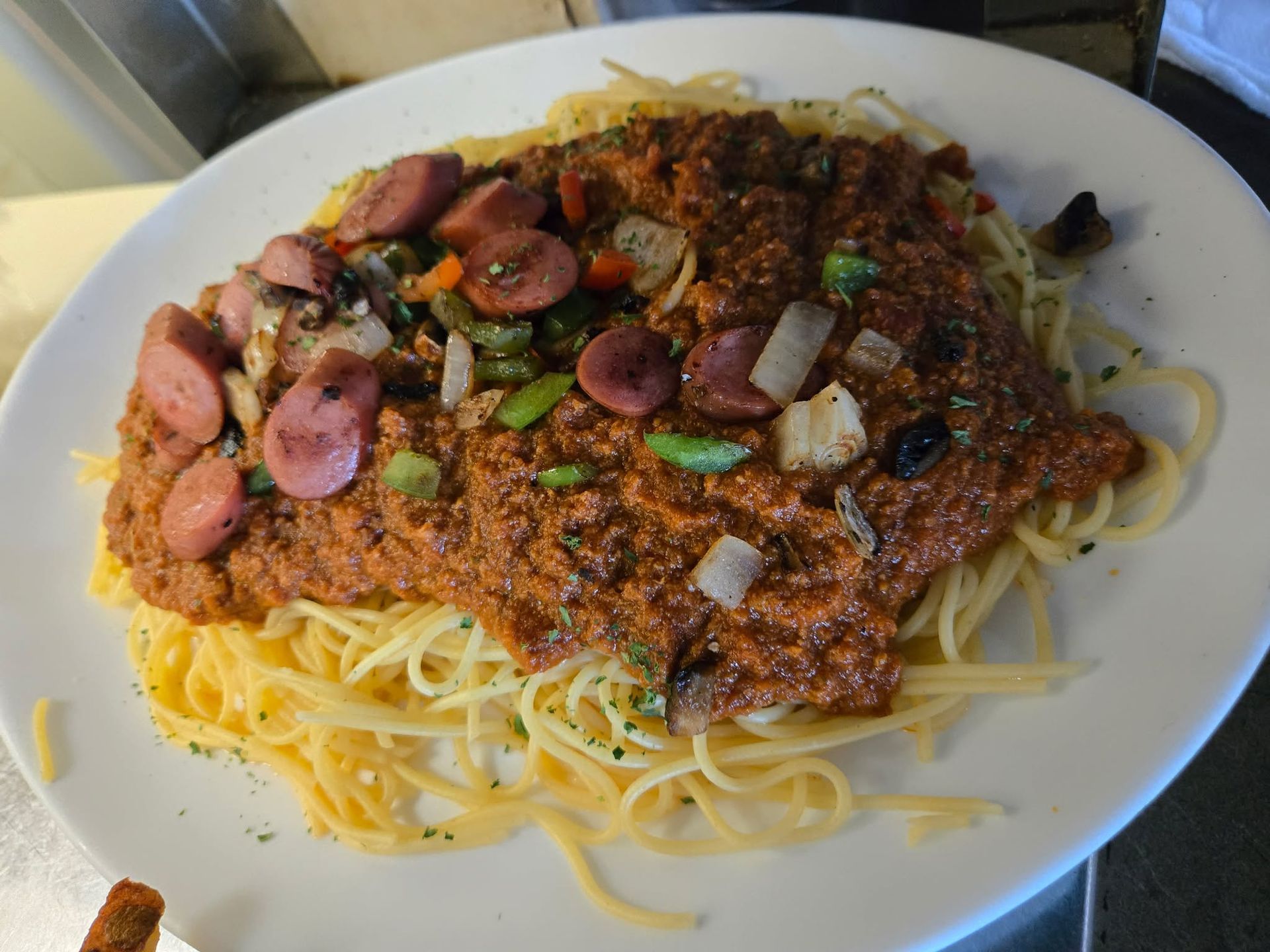 Une assiette de spaghettis nappée d'une sauce à la viande épaisse et rougeâtre, de morceaux de saucisse, d'oignons et de poivrons verts.