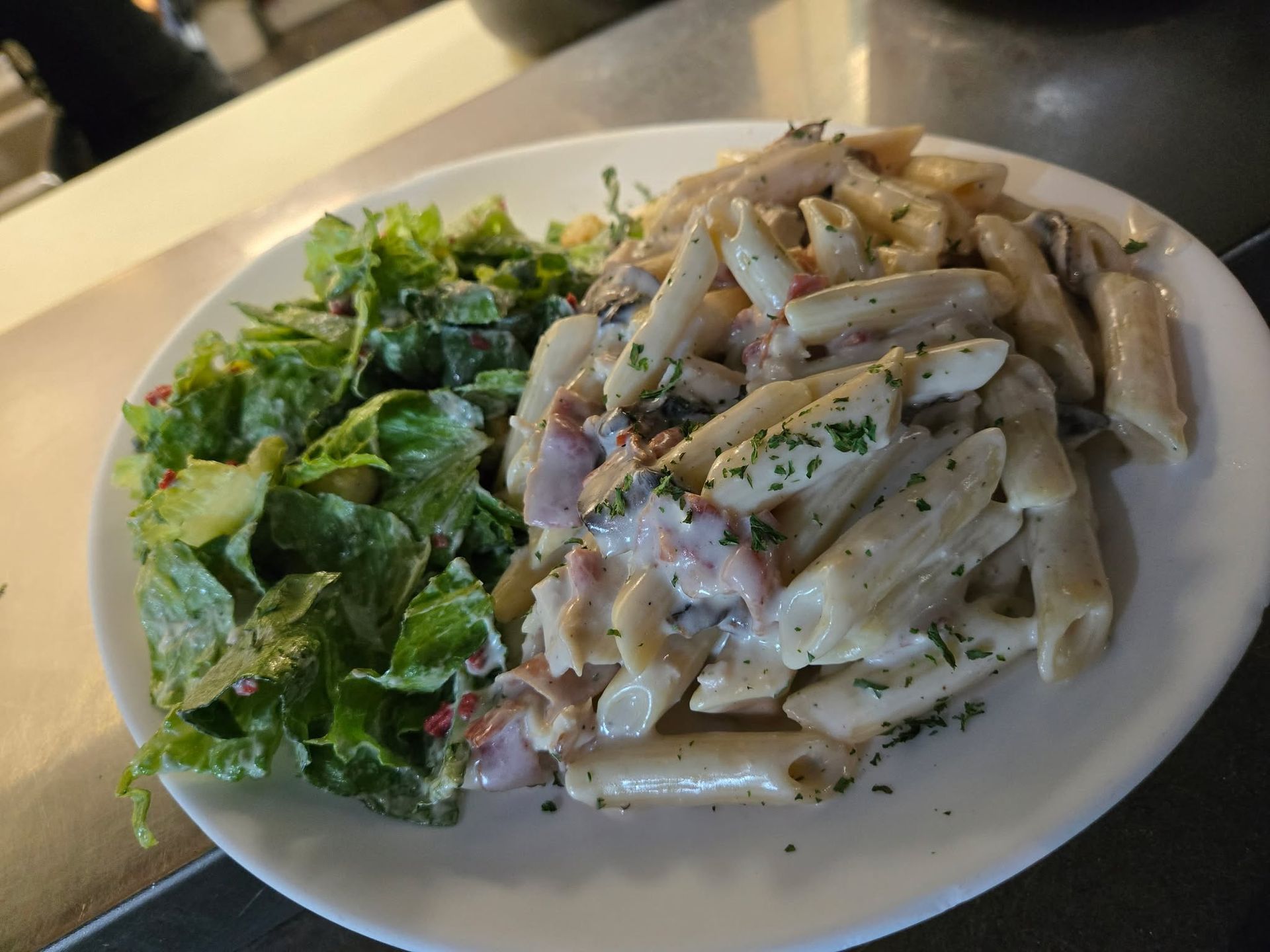 Une assiette blanche contenant des pâtes penne crémeuses au jambon et aux herbes, accompagnées d'une salade verte fraîche.