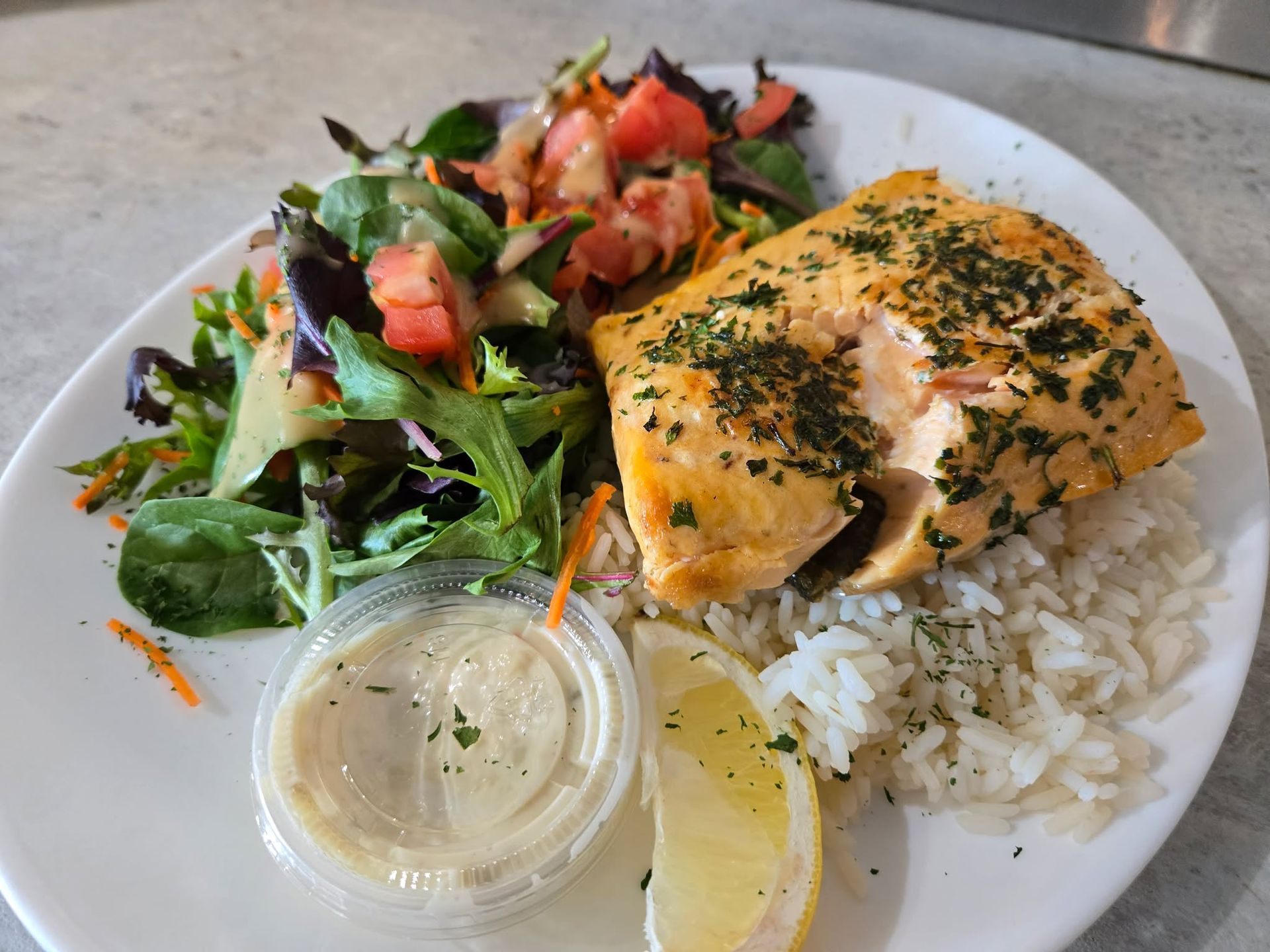 Une assiette composée d'un filet de saumon assaisonné aux herbes, accompagné de riz blanc, d'une salade verte, d'un quartier de citron et de sauce.