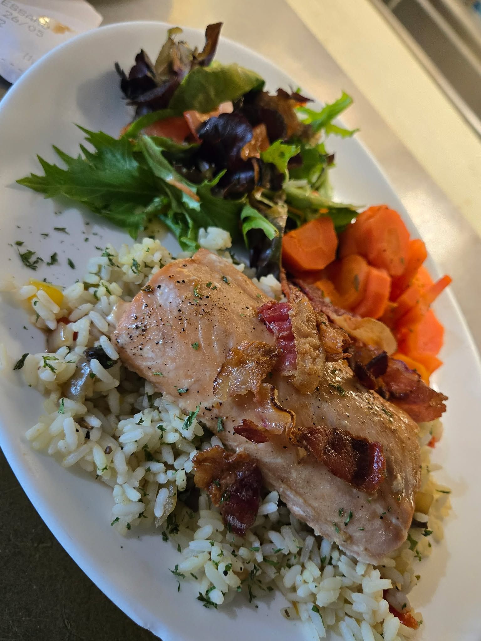 Un plat composé d'un morceau de saumon cuit garni de bacon, servi sur du riz assaisonné avec des carottes et des légumes verts.