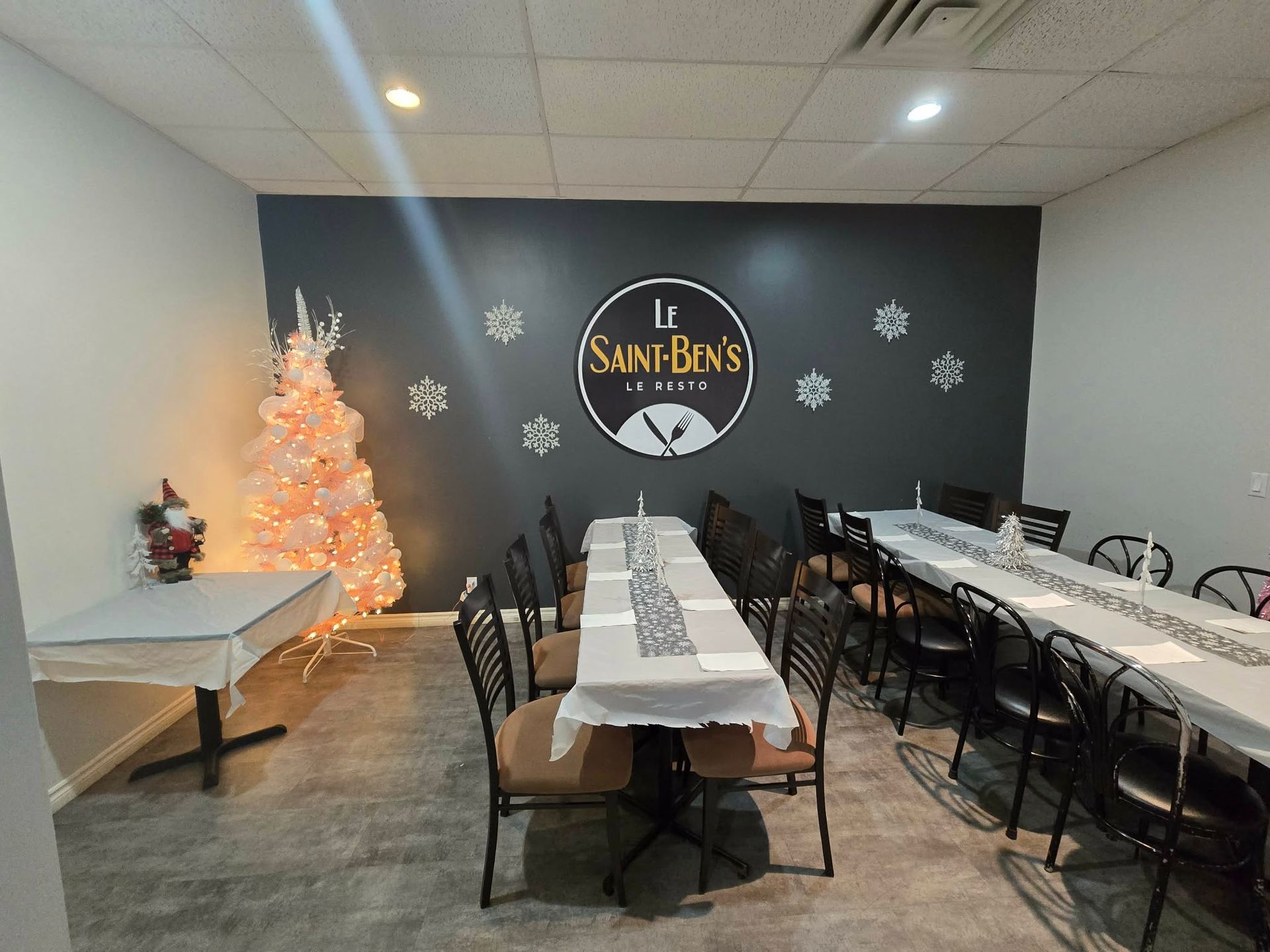 Une salle à manger festive avec de longues tables nappées de blanc, des chaises et un sapin de Noël blanc illuminé sur un mur gris.