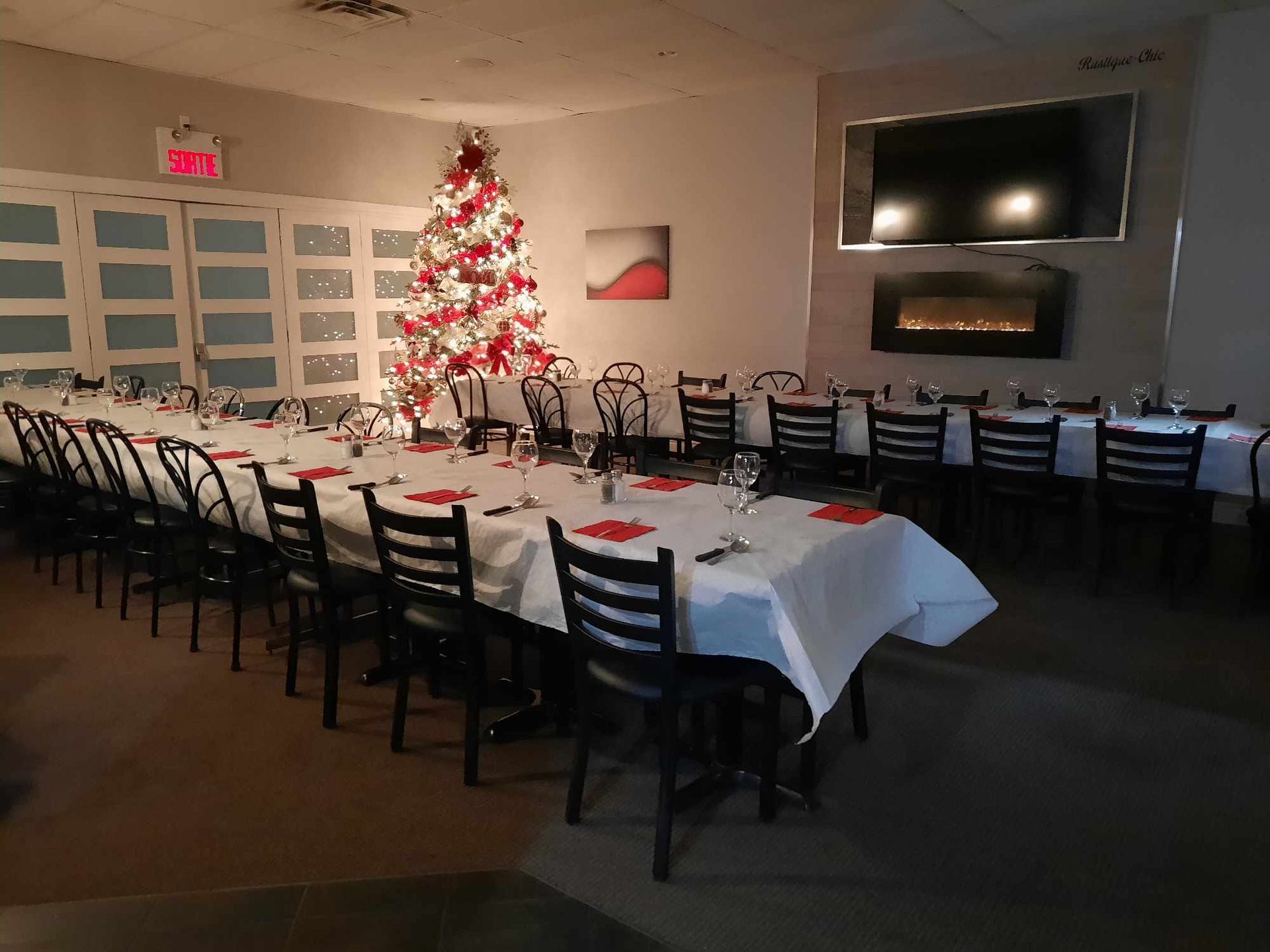 Un sapin de Noël décoré trône dans une salle de banquet où de longues tables recouvertes de nappes blanches sont dressées pour un dîner.