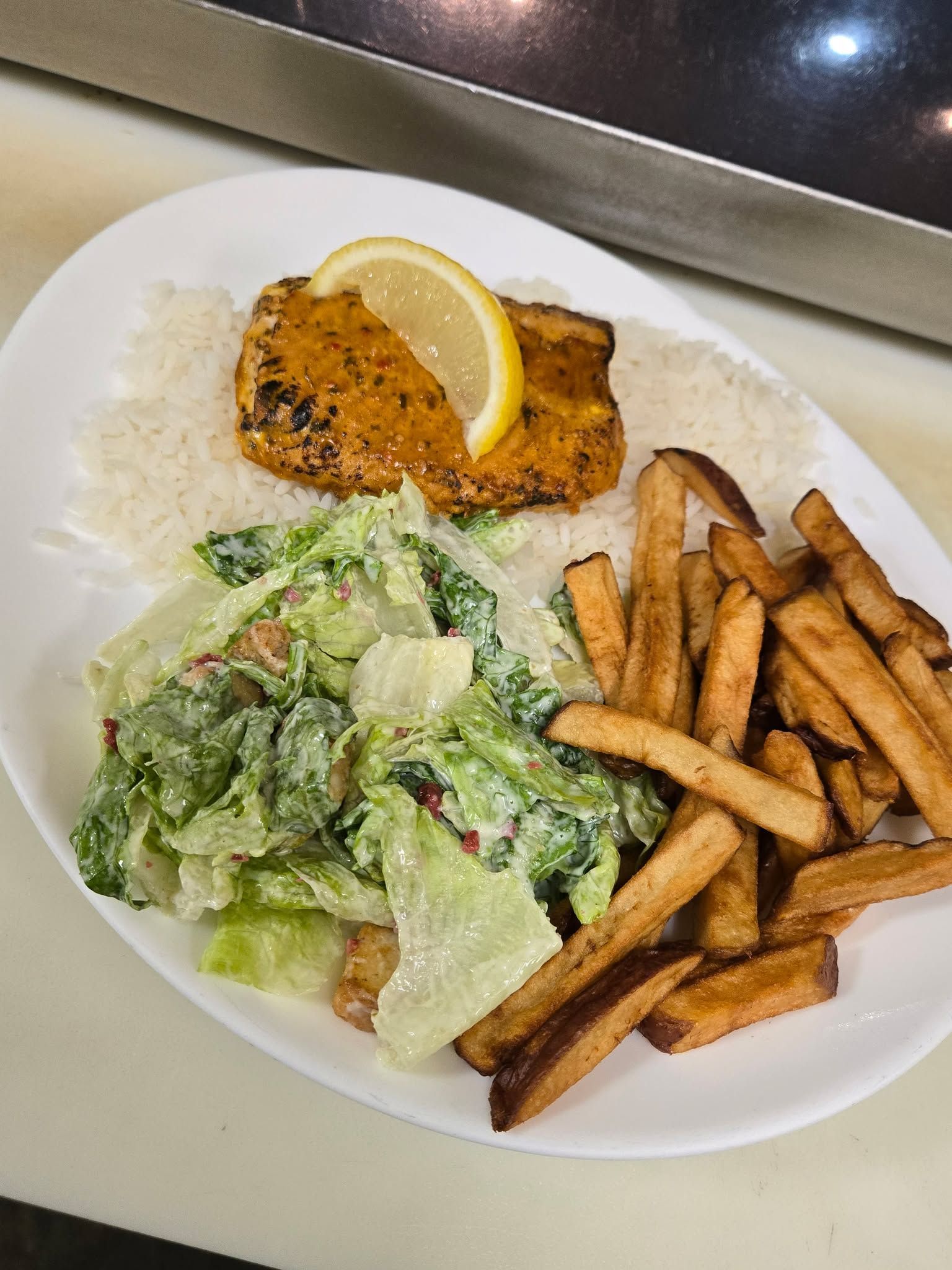 Une assiette blanche contenant un filet de poisson citronné sur du riz, une salade César et une portion de frites.