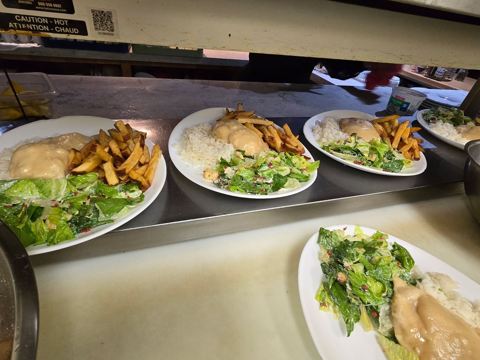 Plusieurs assiettes blanches contenant des salades, des frites et des plats nappés de sauce sont posées sur un plan de travail métallique de cuisine.