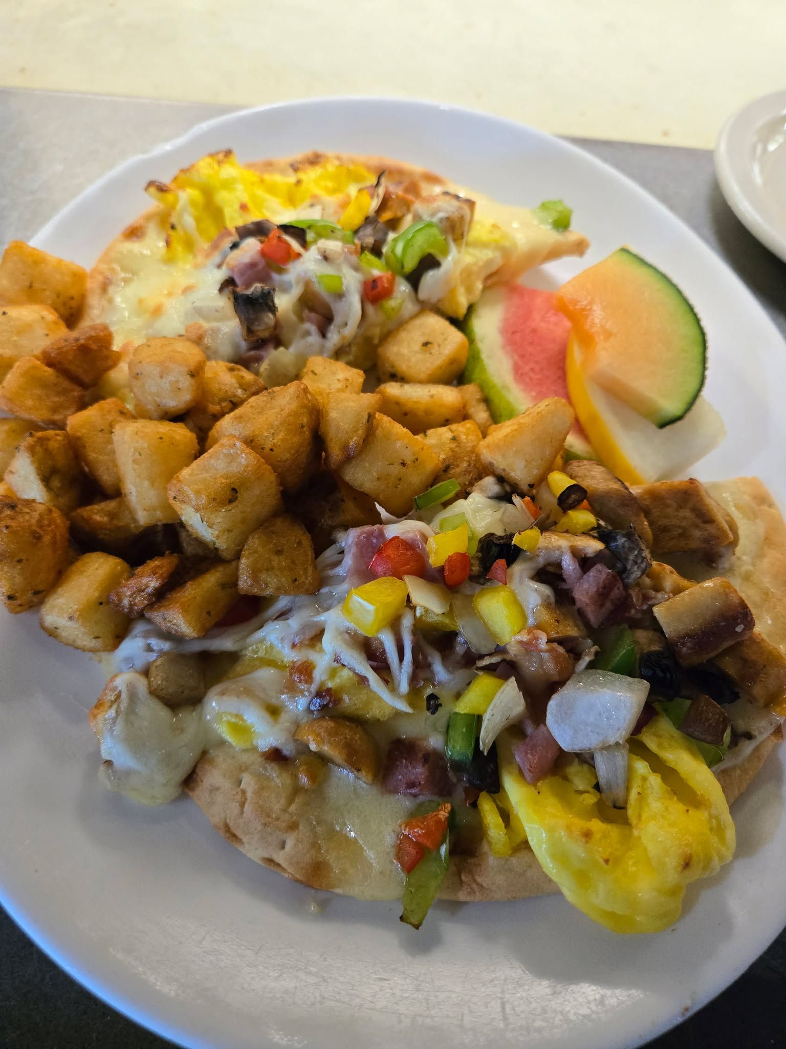 Deux pains pita pour le petit-déjeuner, garnis d'œufs, de poivrons, d'oignons et de viande, servis avec des pommes de terre rissolées et des tranches de fruits.