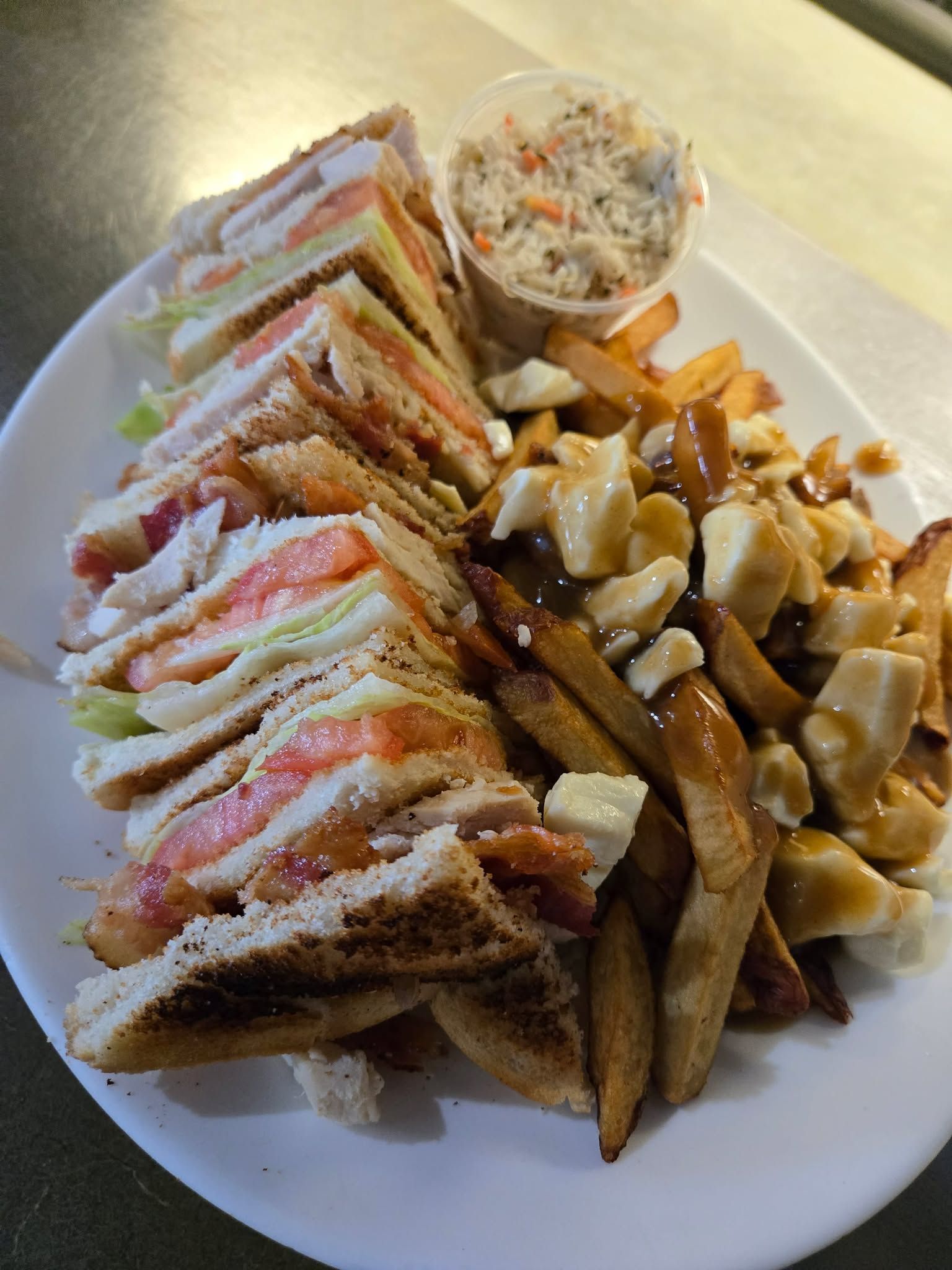 Un club sandwich servi avec de la poutine et une salade de chou sur une assiette blanche.