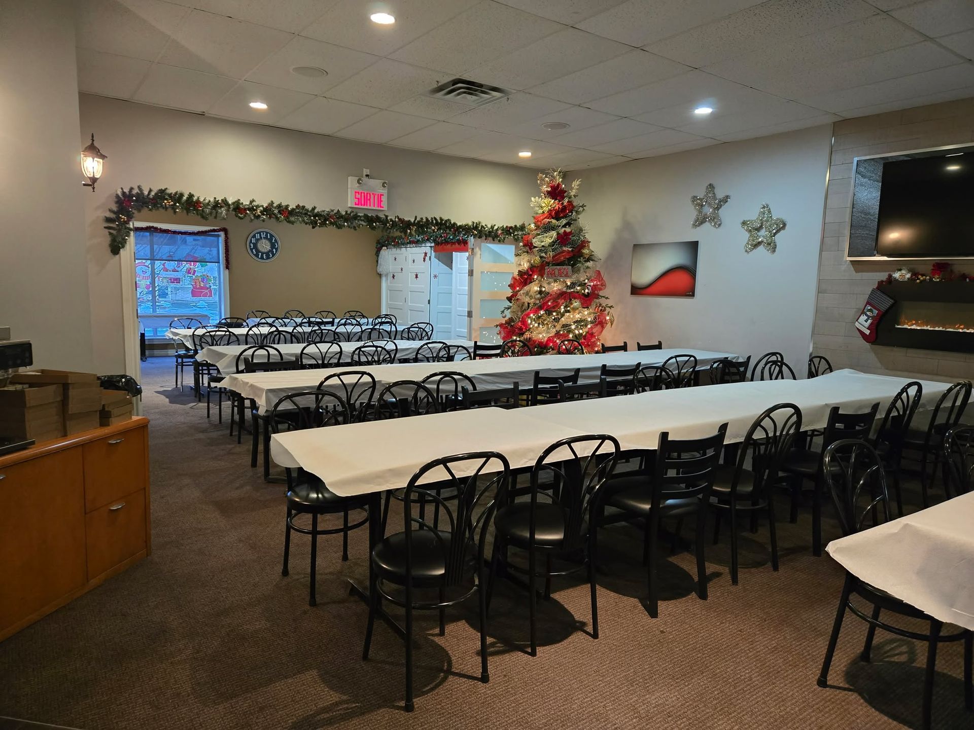 Une salle à manger préparée pour une fête, avec de longues tables recouvertes de nappes blanches, des chaises noires et un arbre décoré.