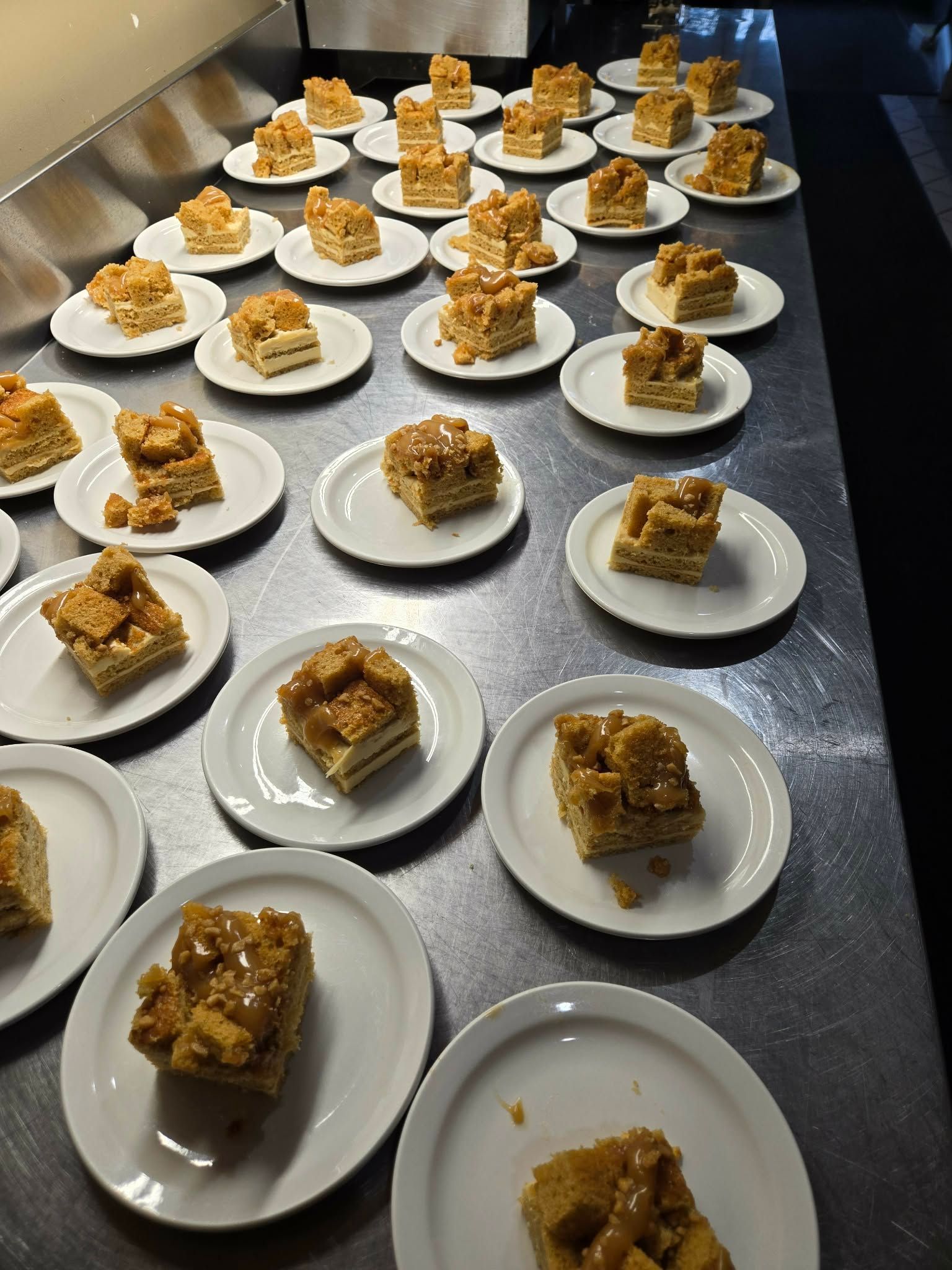 De nombreuses petites assiettes surmontées de morceaux de gâteau carrés sont disposées sur un long plan de travail métallique de cuisine professionnelle.