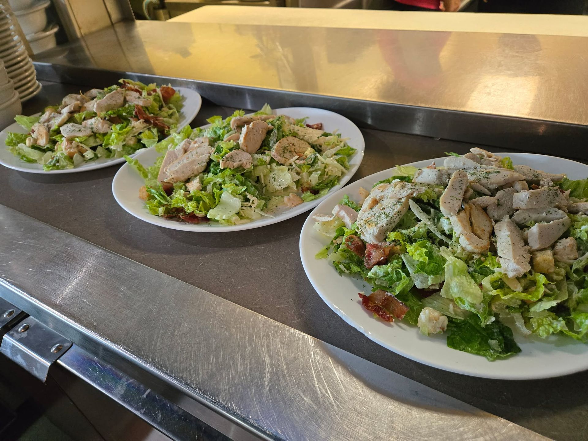 Trois assiettes blanches de salade César au poulet sont disposées sur un comptoir de service en acier inoxydable.