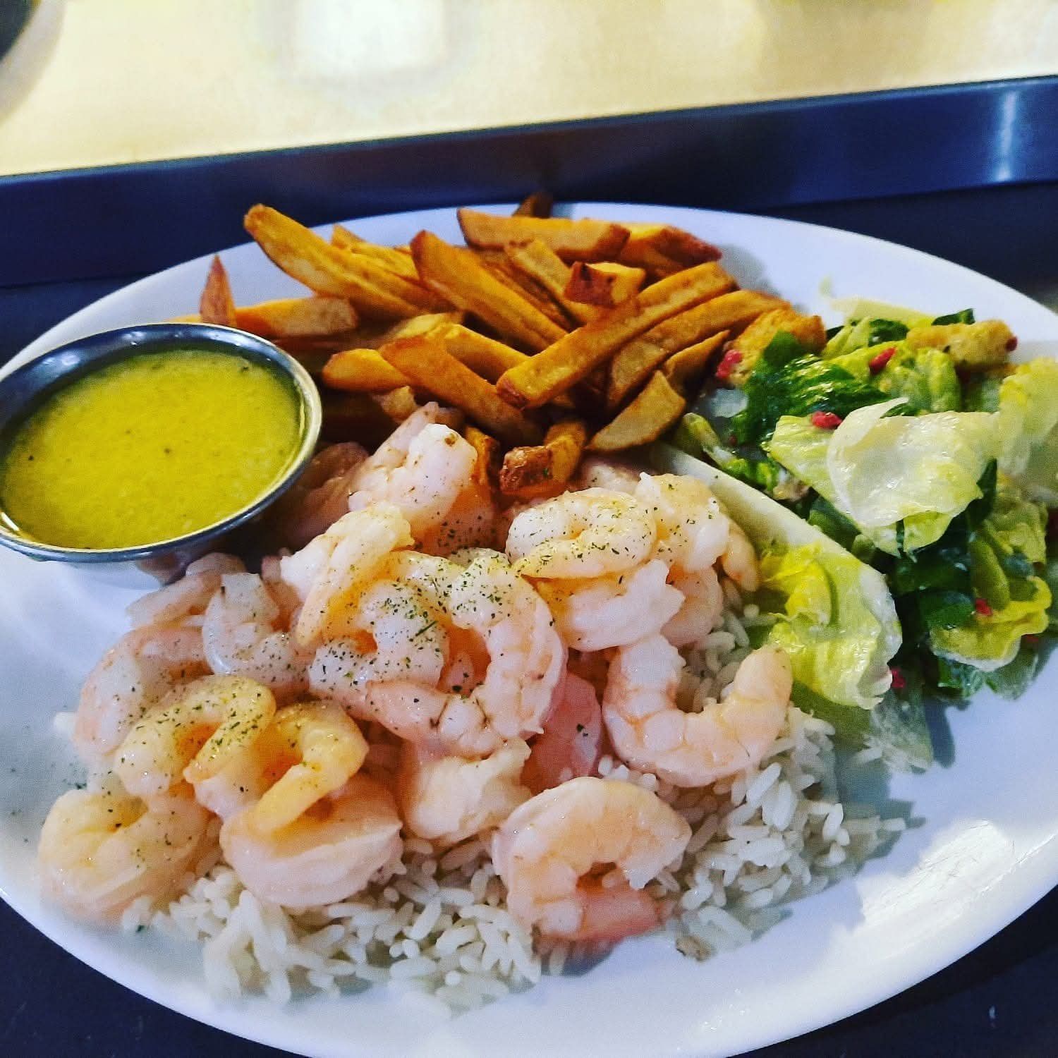 Une assiette blanche présente des crevettes assaisonnées sur du riz blanc, servies avec des frites, une salade verte et une petite coupelle de sauce.