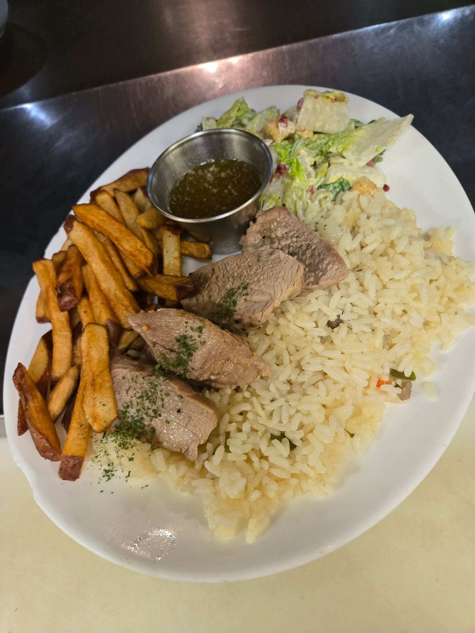 Une assiette composée de viande assaisonnée, de riz, de frites, d'une petite coupelle de sauce brune et d'une salade d'accompagnement.