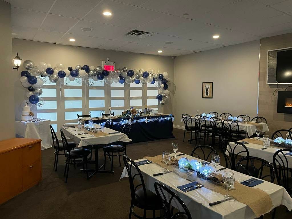Salle de banquet décorée avec tables dressées, chaises noires et guirlande de ballons bleus et blancs le long des fenêtres