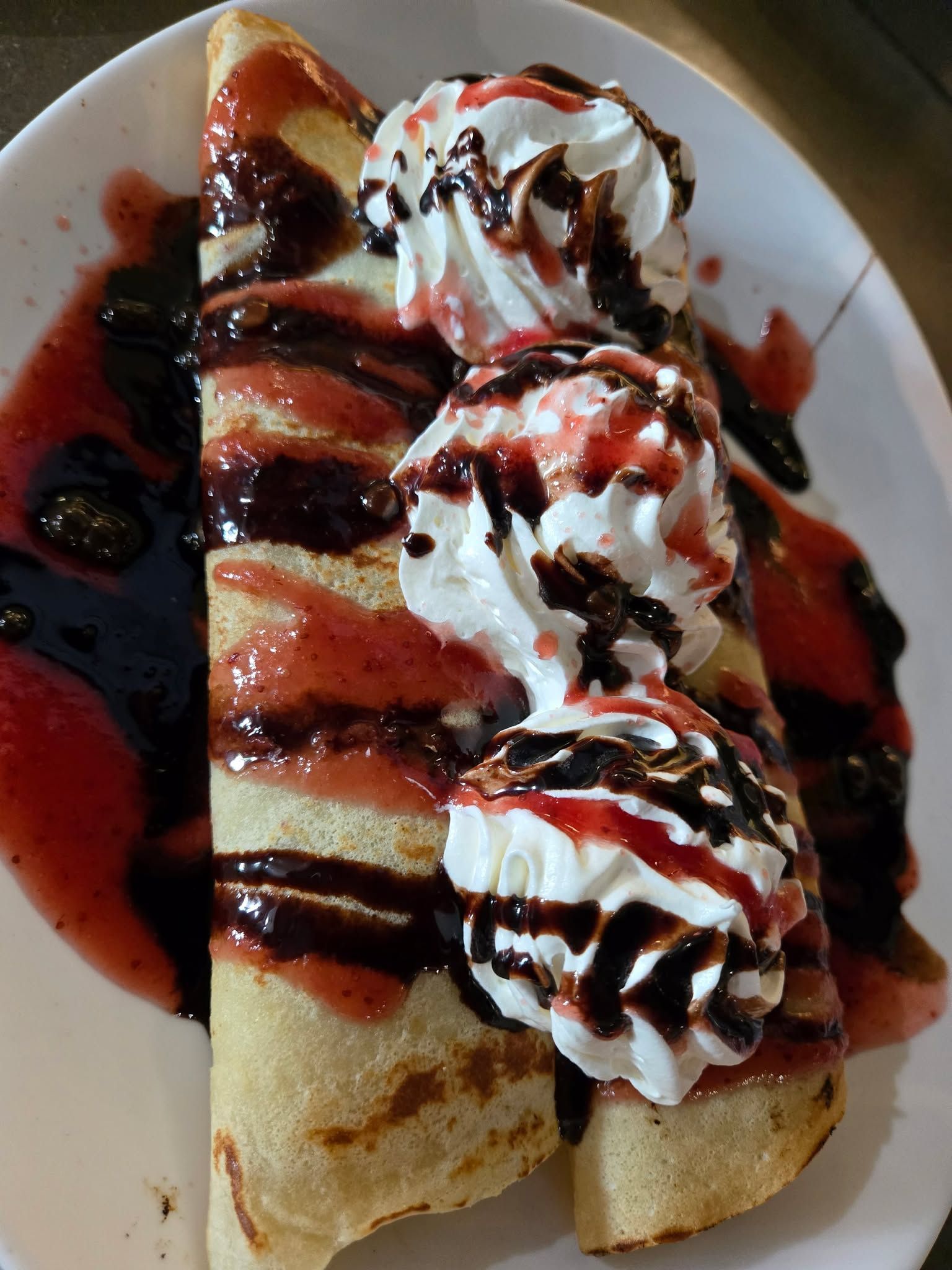 Crêpes roulées sur une assiette blanche, arrosées de sauces au chocolat et aux fraises, et garnies de crème fouettée.