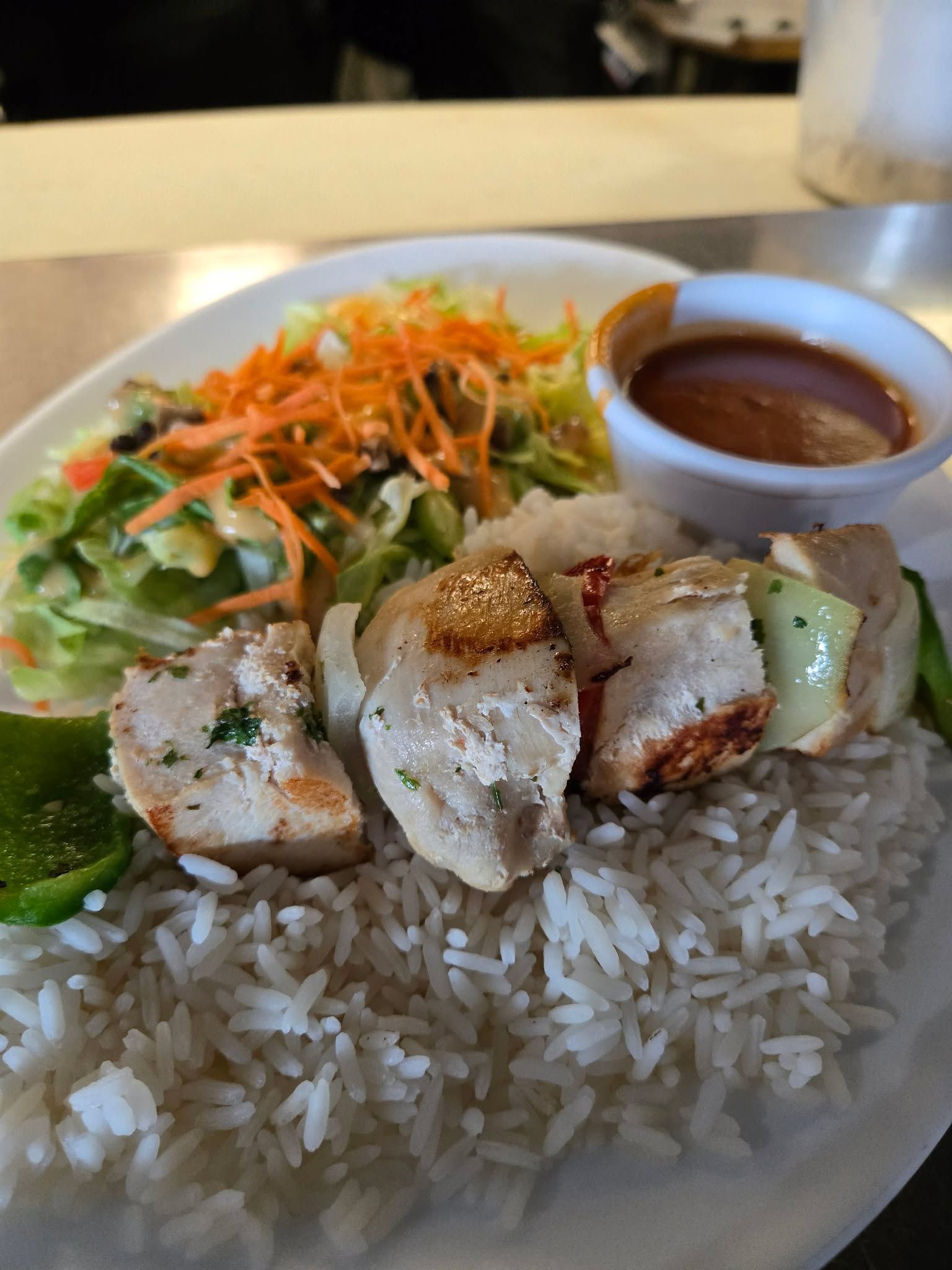 Brochettes de poulet servies avec du riz blanc, une salade d'accompagnement avec des carottes râpées et un petit ramequin de sauce.