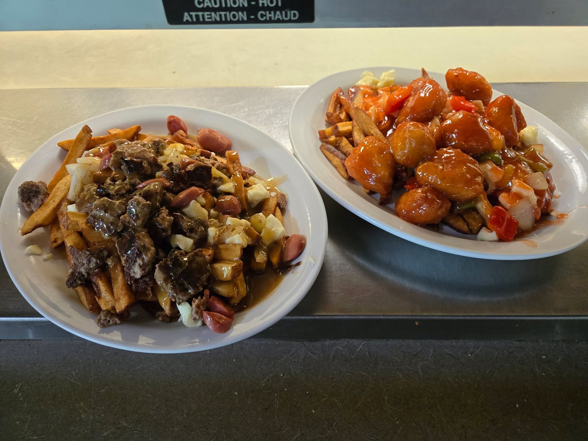 Deux assiettes blanches de poutine, l'une garnie de viande et de saucisses, l'autre de poulet pané et nappé de sauce, posées sur un comptoir.