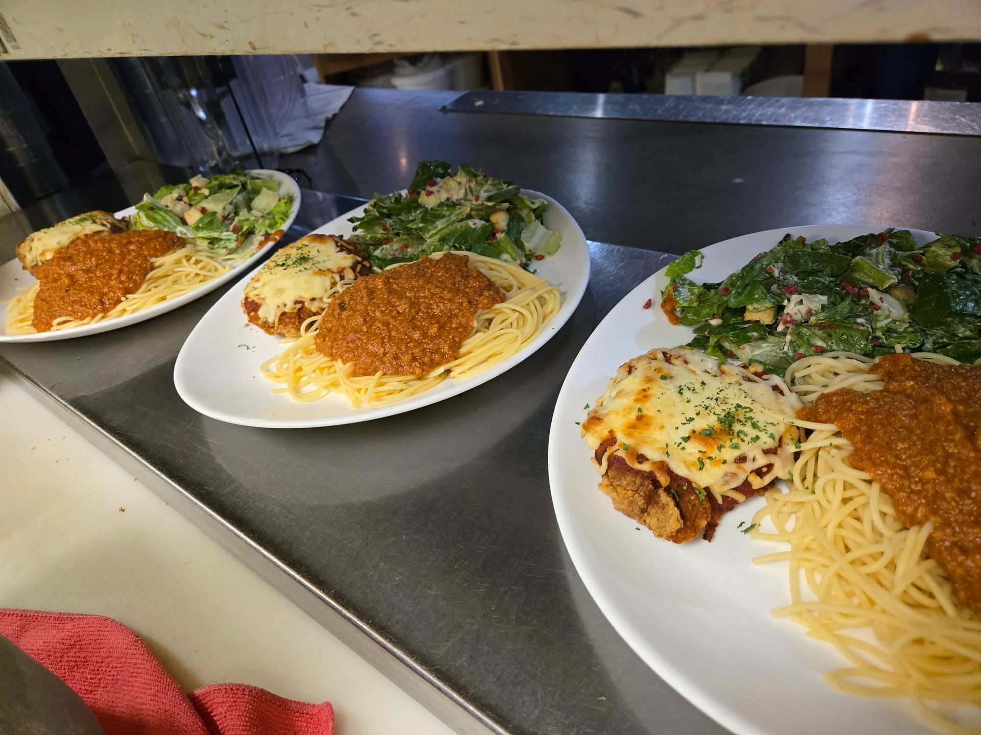 Trois assiettes sur un comptoir en métal, chacune contenant des spaghettis à la sauce bolognaise, une escalope panée au fromage fondu et de la salade.